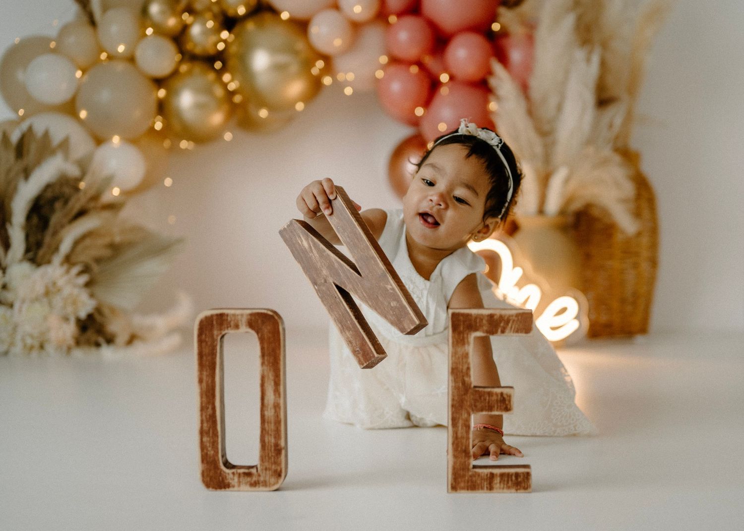 Baby girl in white romper plays with wooden ONE letters at boho first birthday cake smash session.