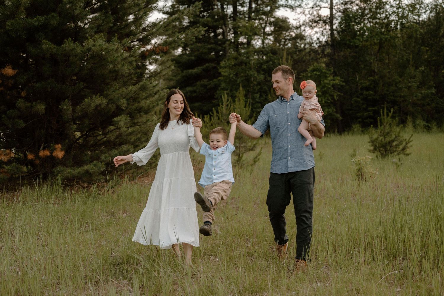 A happy family swings their toddler while walking through a lush green meadow.
