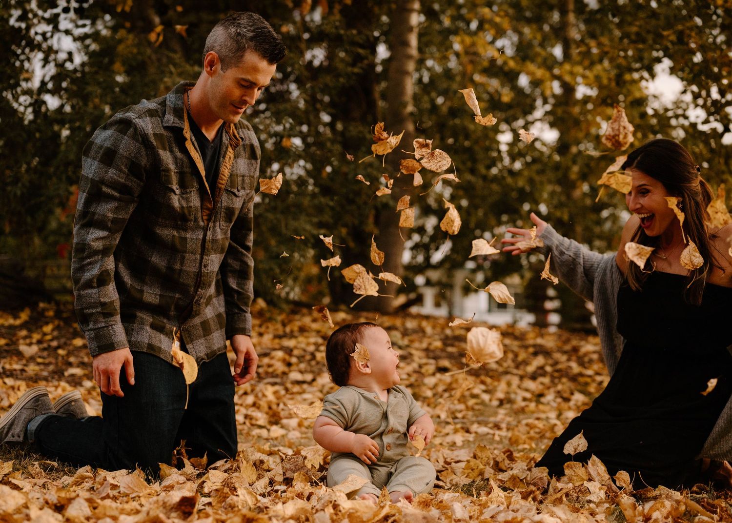 Family playing in autumn leaves with baby sitting surrounded by golden fall foliage outdoors.