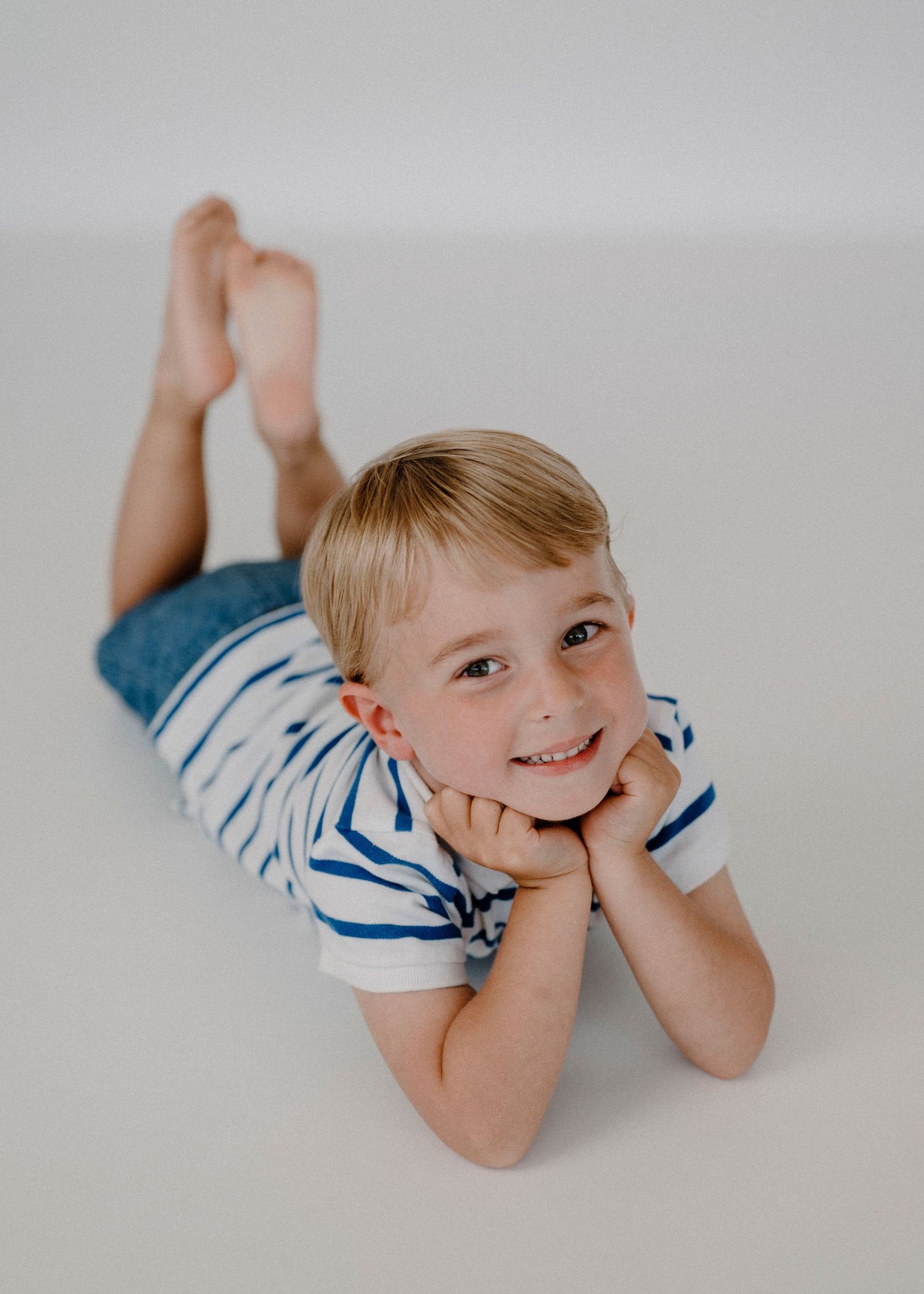 Cute blonde boy in striped shirt lying on stomach, smiling with chin resting on hands.
