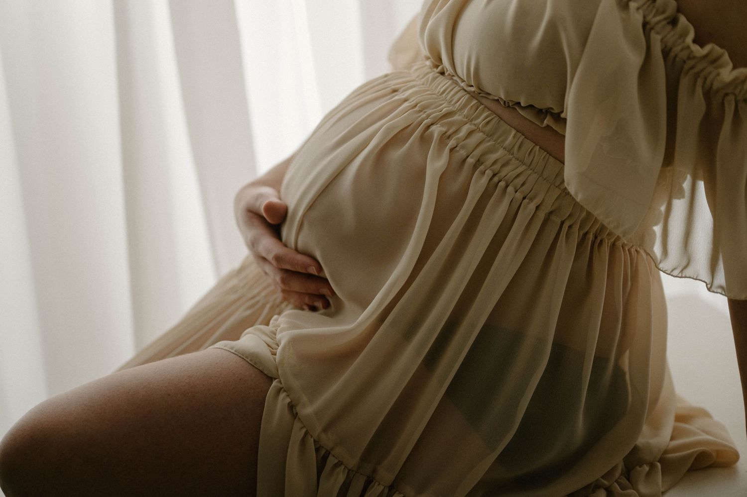 Close-up of a pregnant woman in a ruffled cream dress gently cradling her baby bump near sheer curtains.