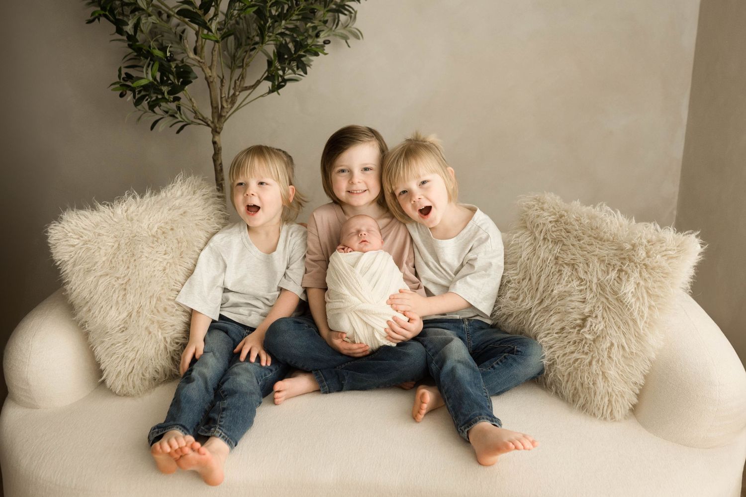 Three happy siblings sit on a white couch, smiling and laughing while holding a sleeping newborn baby wrapped in white.