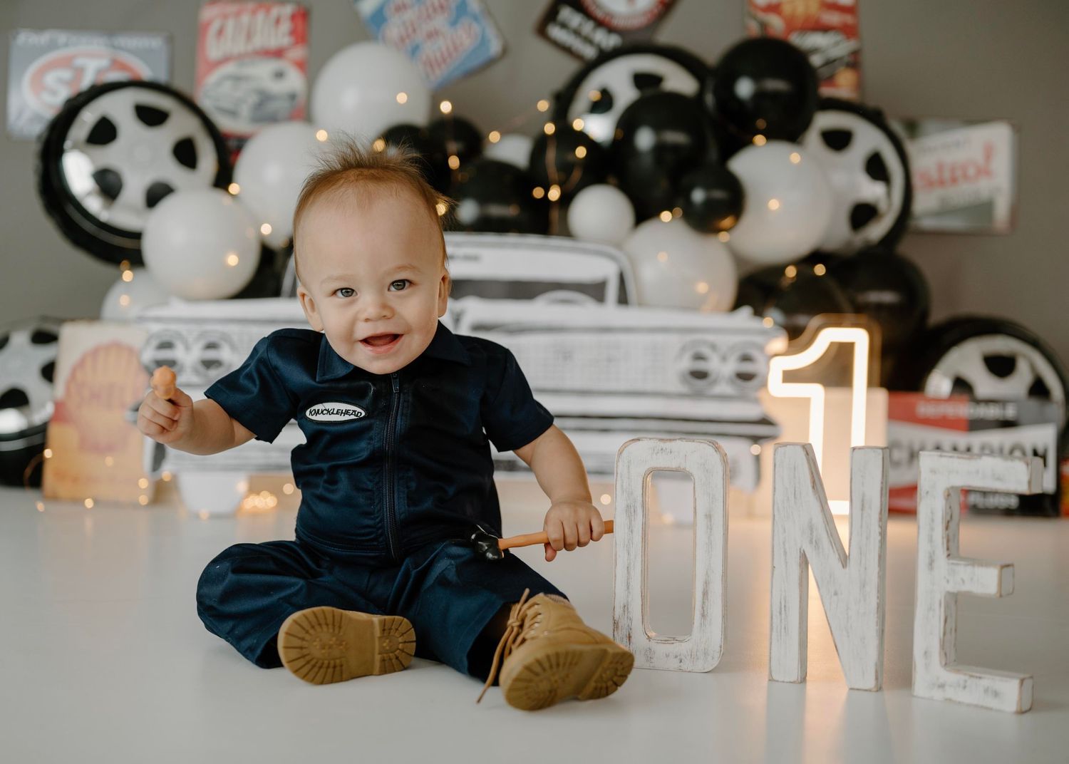 Smiling baby boy in mechanic jumpsuit poses with illuminated ONE sign at garage-themed first birthday shoot.