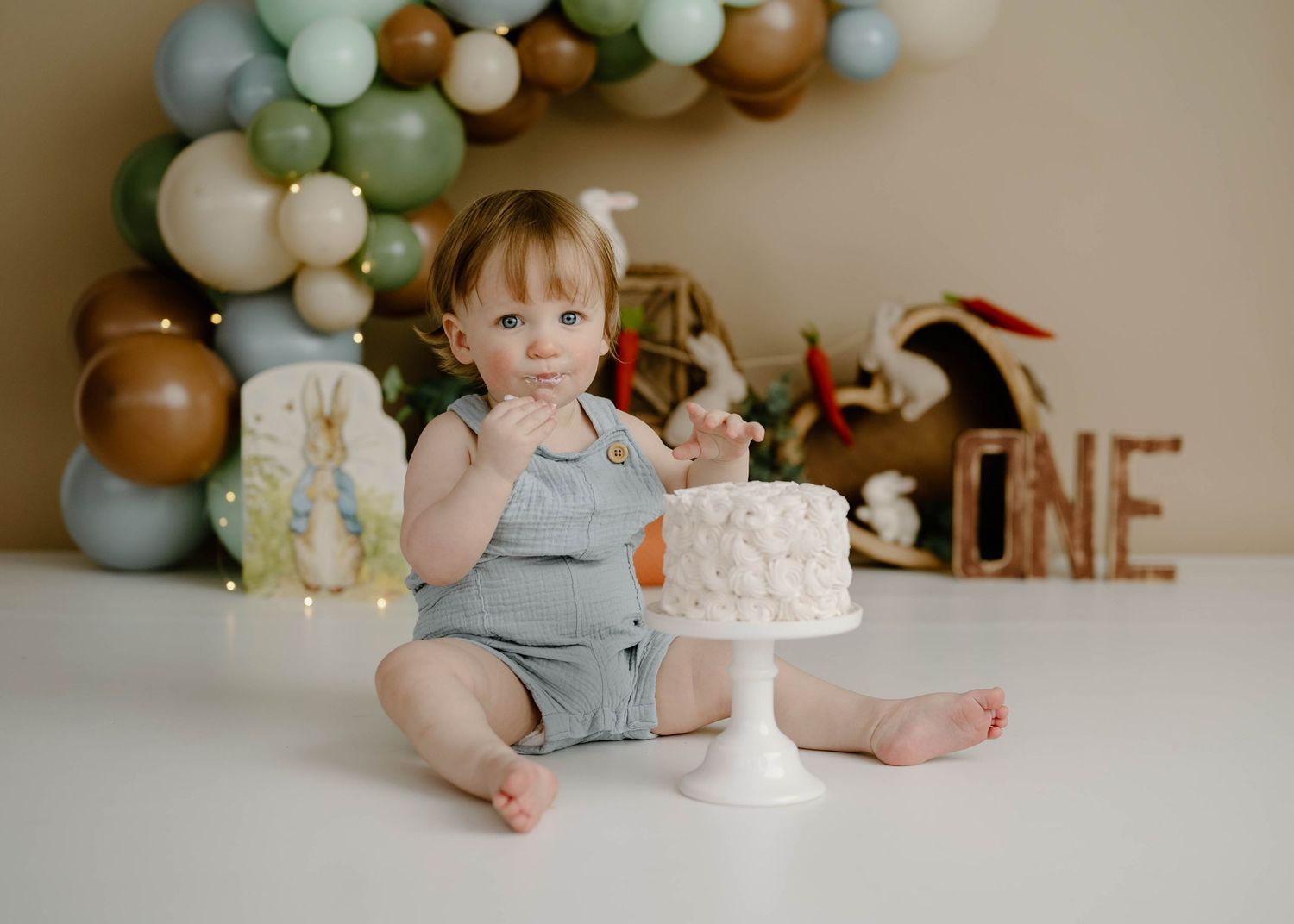 Baby boy eating cake at Peter Rabbit themed first birthday cake smash with green balloons.