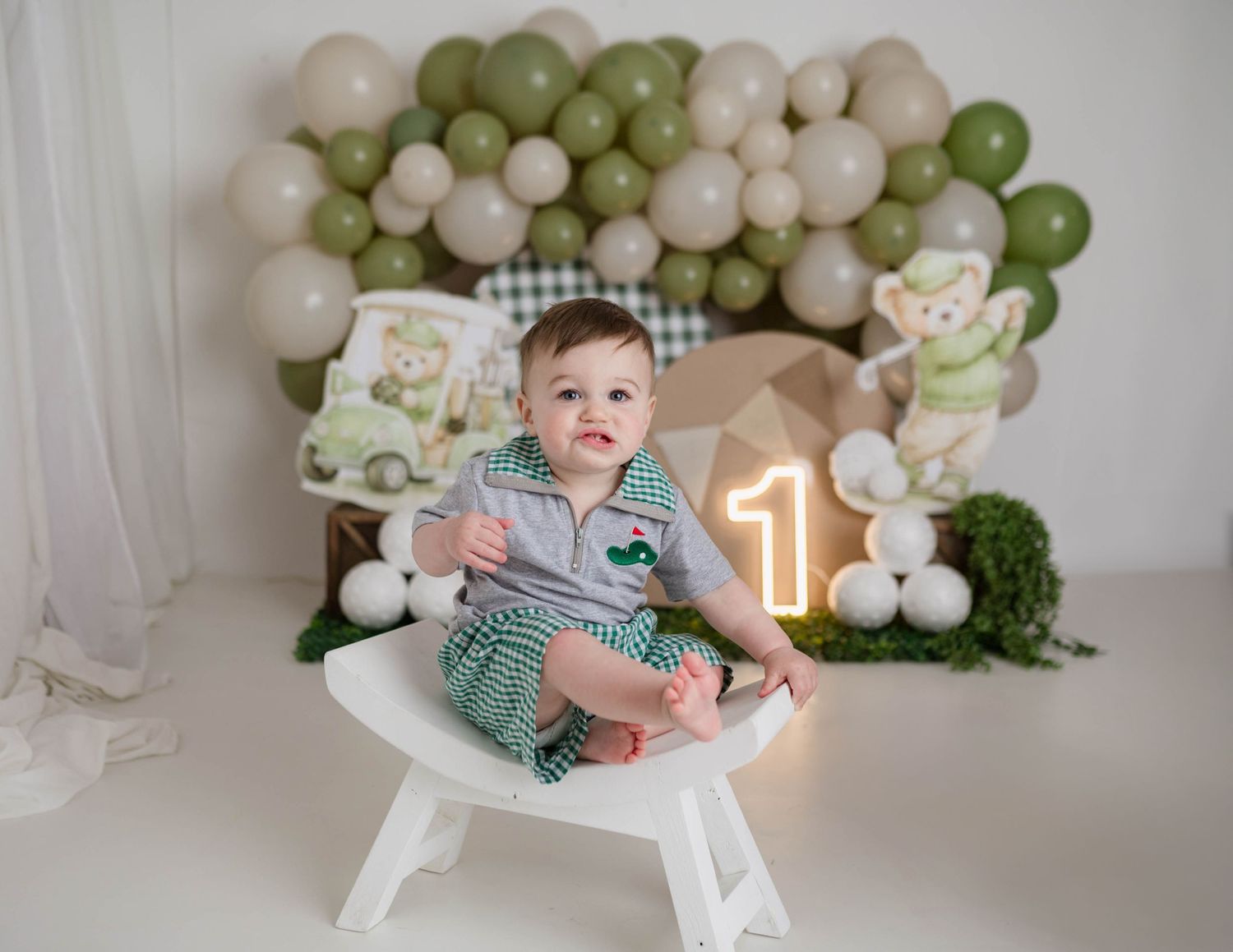 Smiling baby boy sits on white chair at golf-themed first birthday setup with green and beige balloon garland.