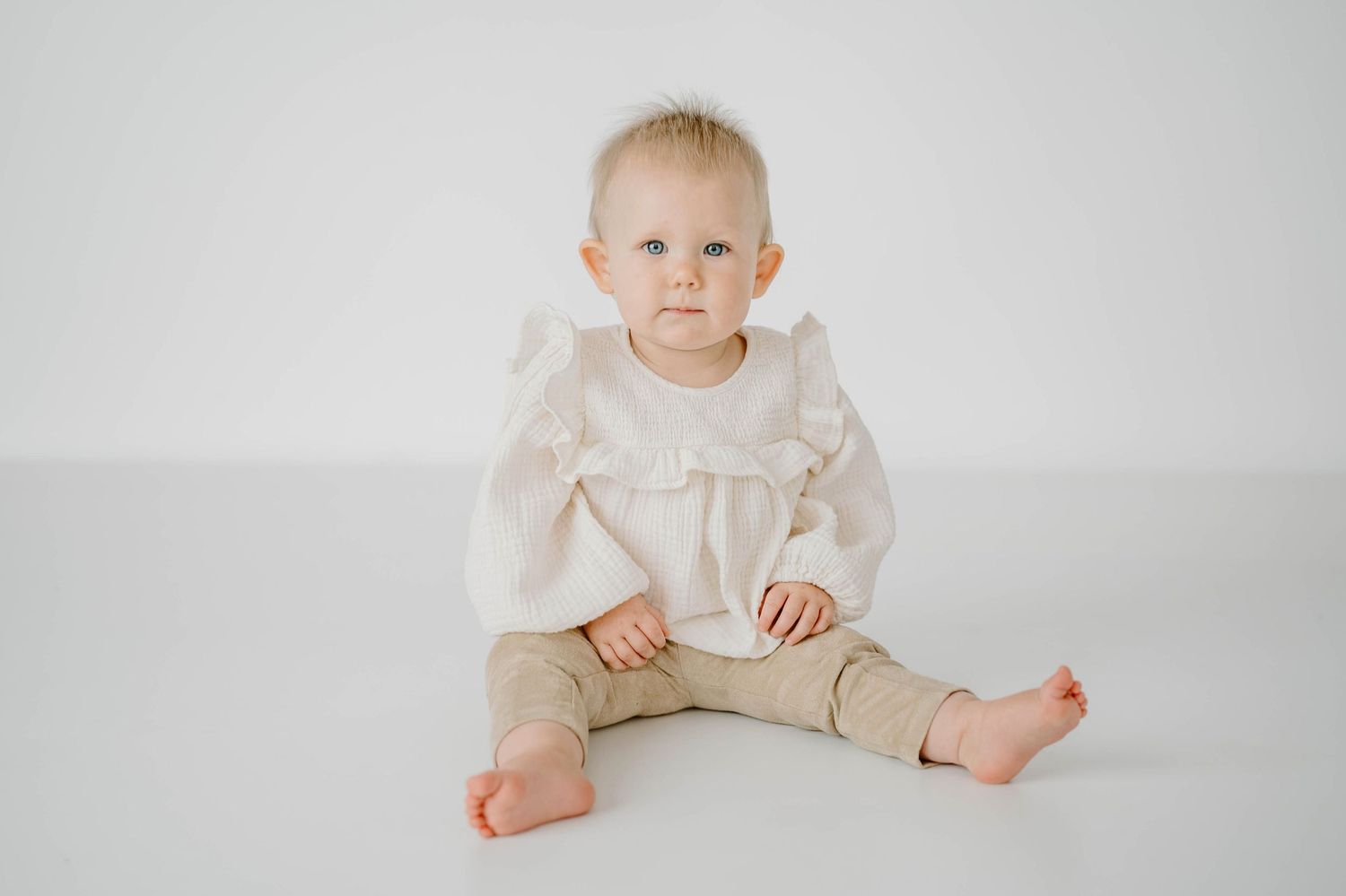 Adorable blue-eyed baby in white ruffle blouse and tan pants sitting on studio floor.