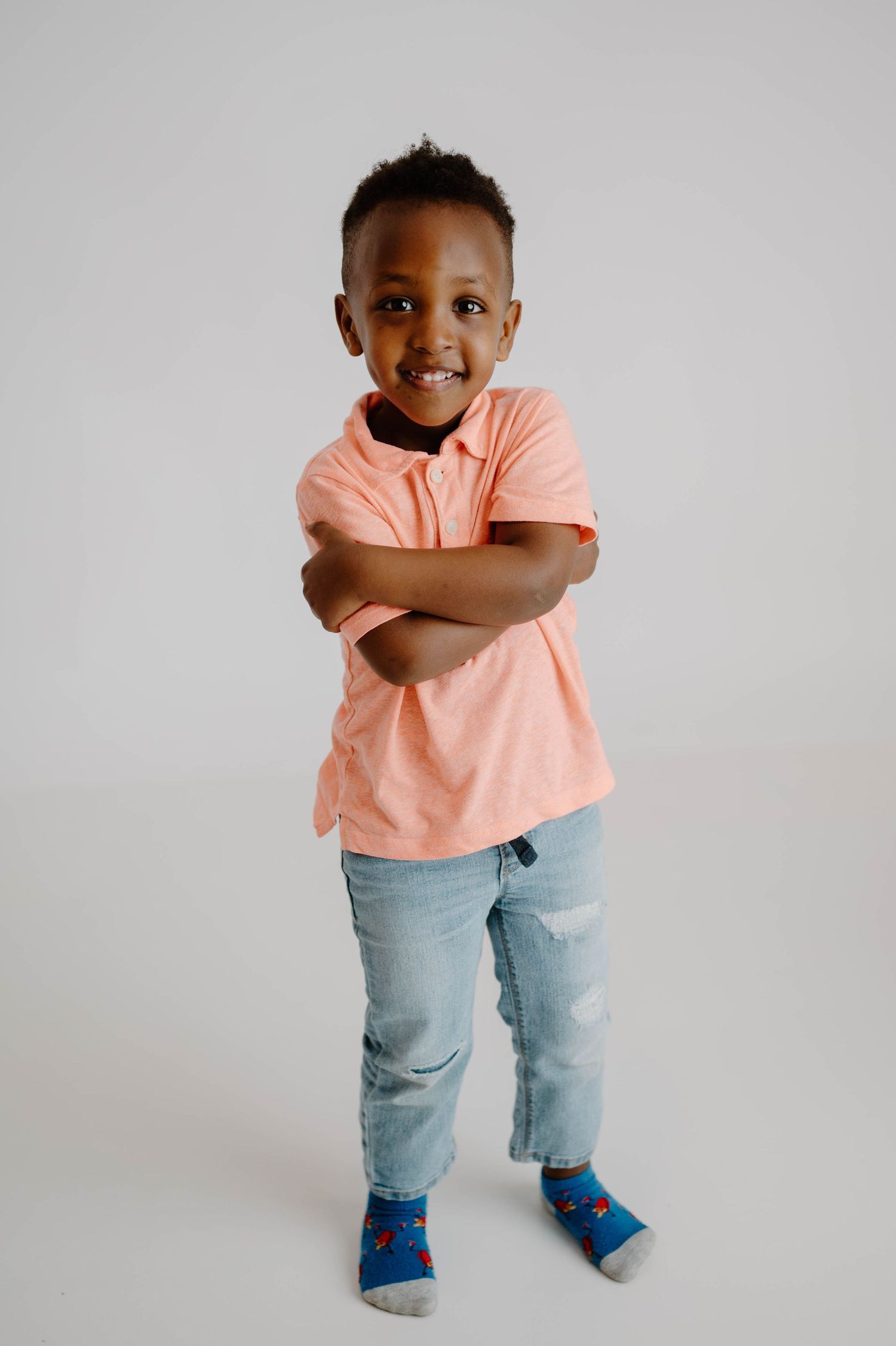 Young Black boy in a pink polo shirt and jeans standing with arms crossed, smiling confidently.