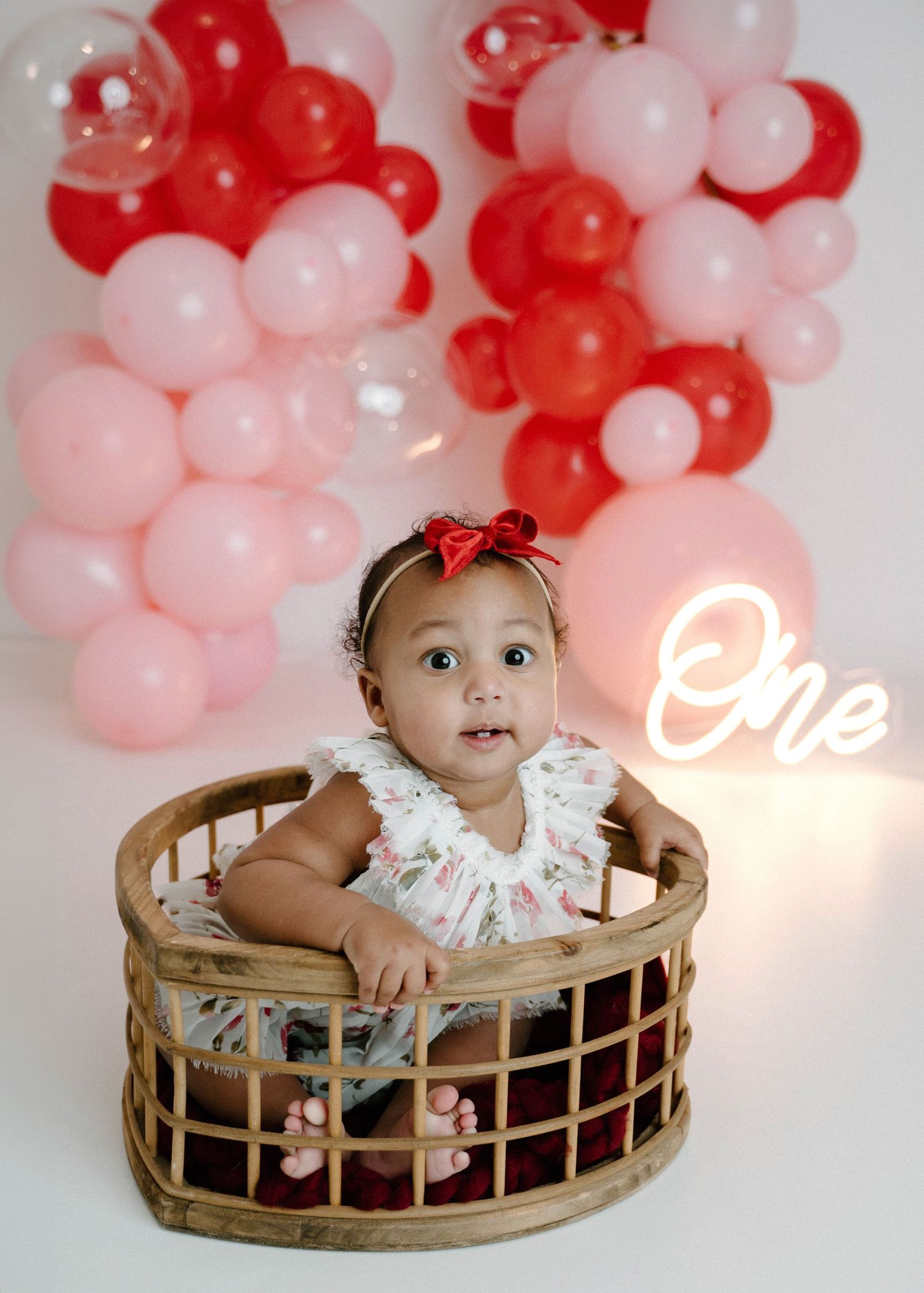Adorable baby girl in floral dress sits in wicker basket with pink red balloon backdrop.