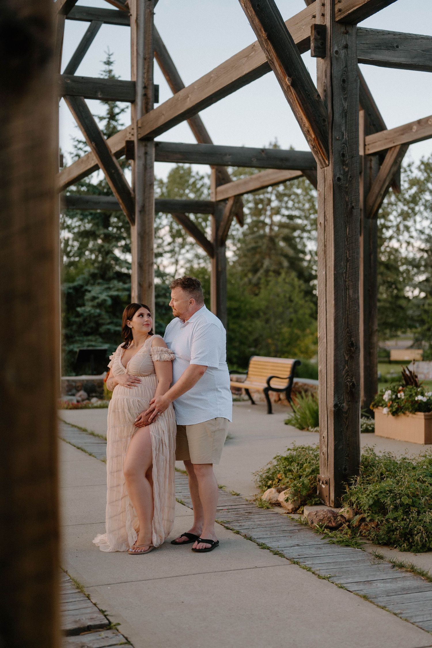 Couple poses under rustic wooden pergola during golden hour maternity photoshoot in lush garden setting.