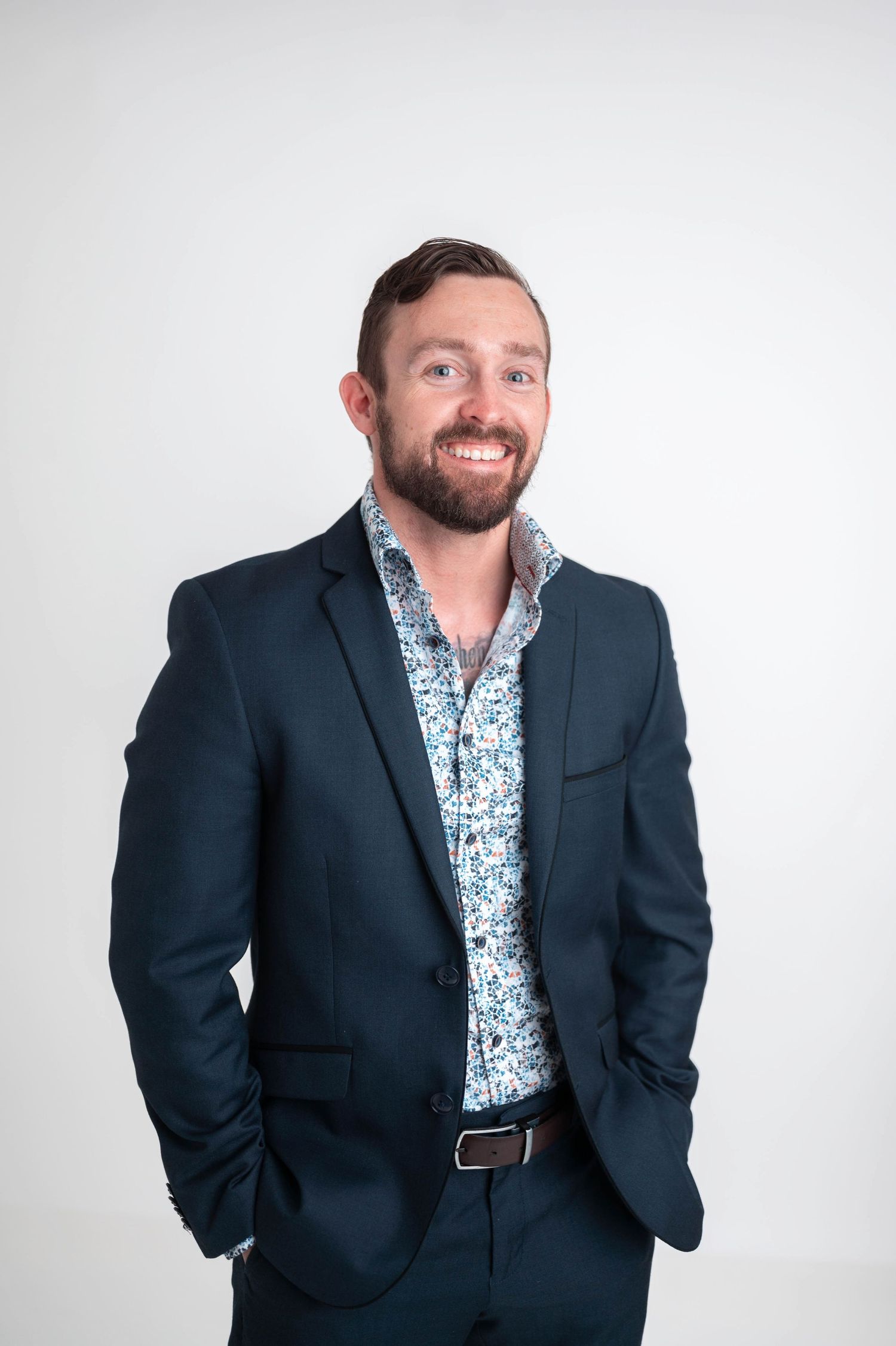 Smiling bearded man in navy suit and floral shirt posing confidently with hands in pockets against white background.
