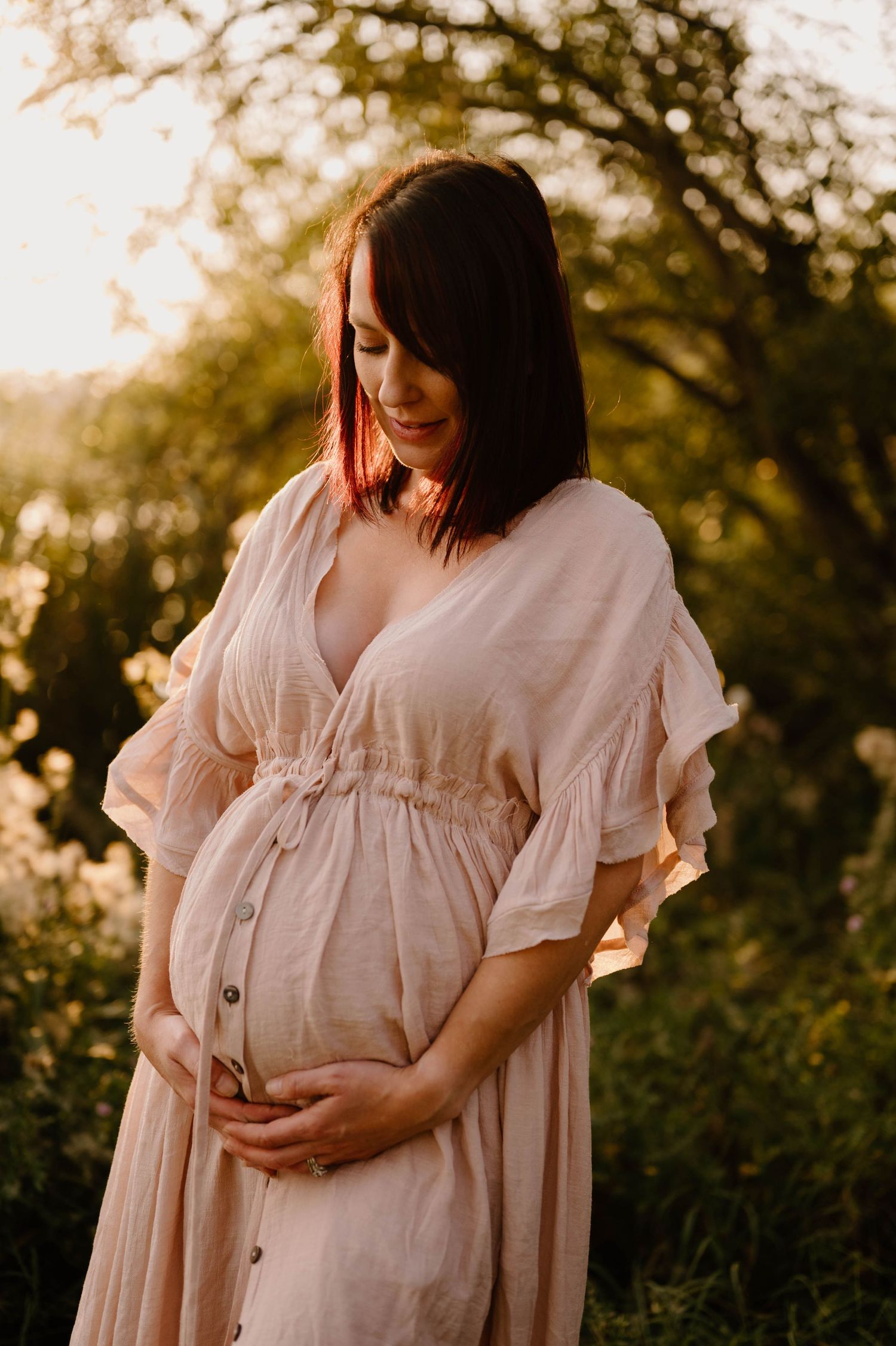 Pregnant woman in blush linen dress holding baby bump outdoors at golden hour.
