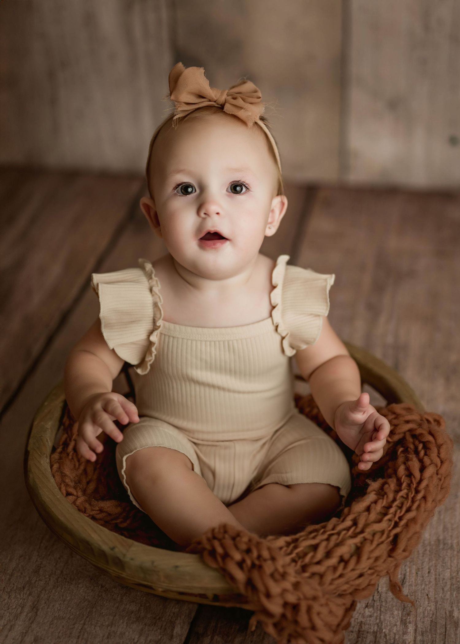 Adorable baby girl in cream ruffle romper and bow headband sitting in wooden bowl prop for studio portrait.