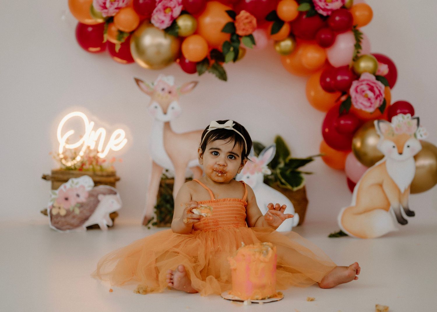 Baby girl in orange tutu dress celebrating first birthday with cake smash and woodland decor.
