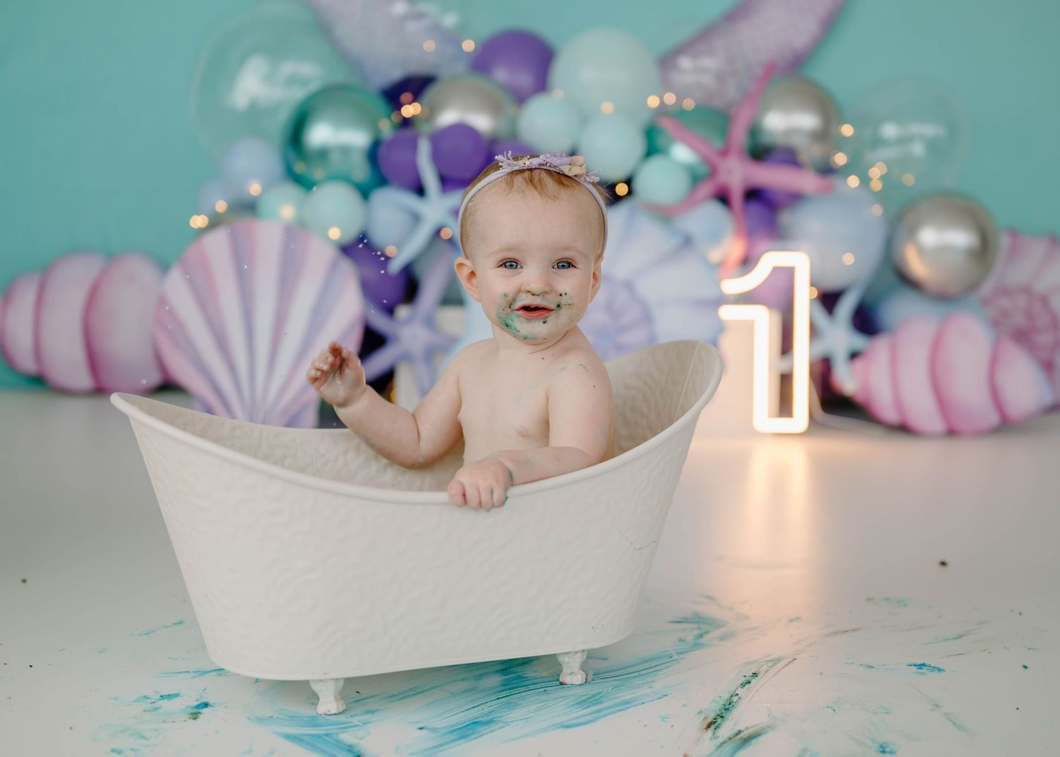 Smiling baby girl sits in white mini bathtub during mermaid first birthday photo session with balloon backdrop.