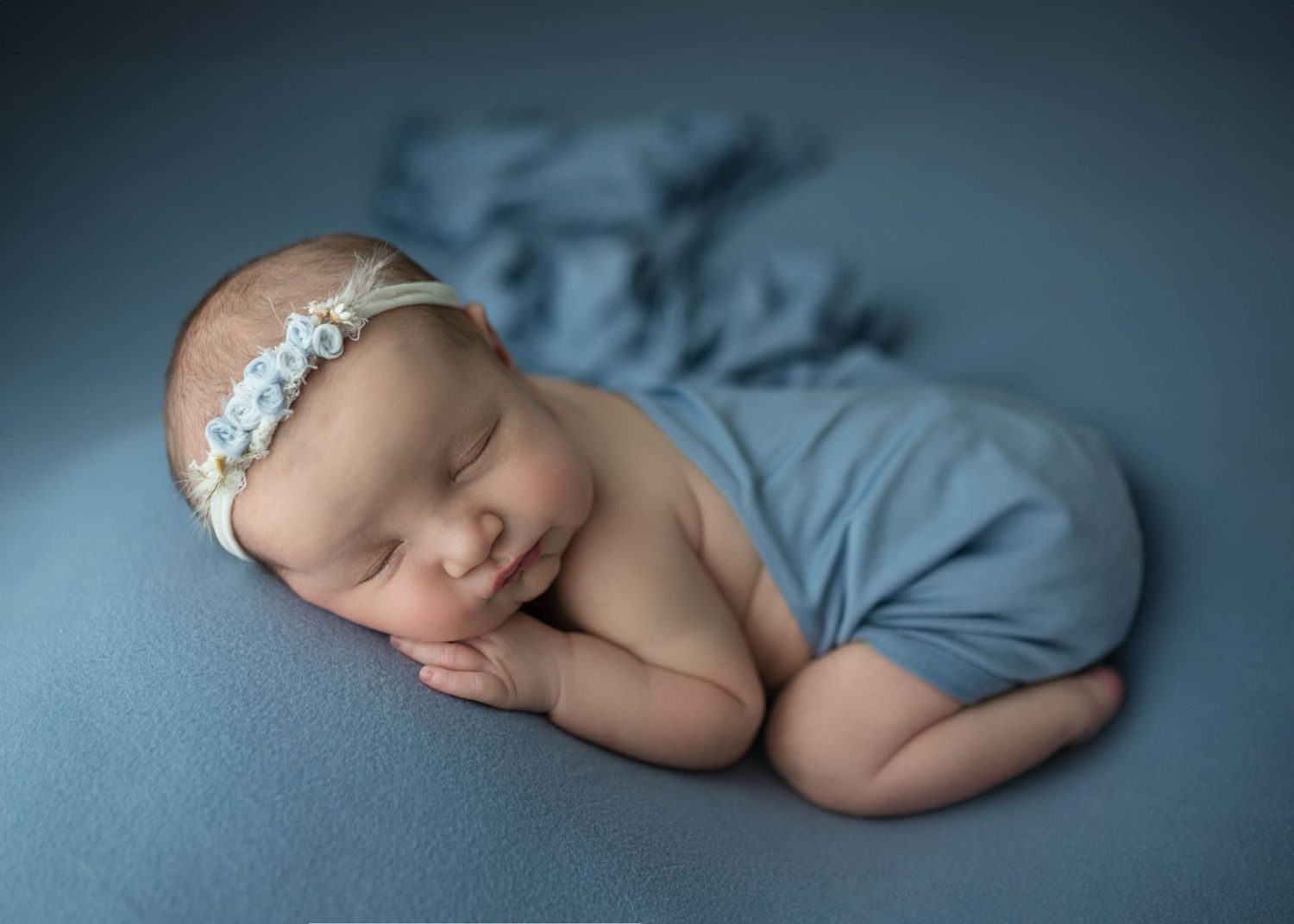 Sleeping newborn baby girl wrapped in blue swaddle with floral headband on soft blue backdrop.