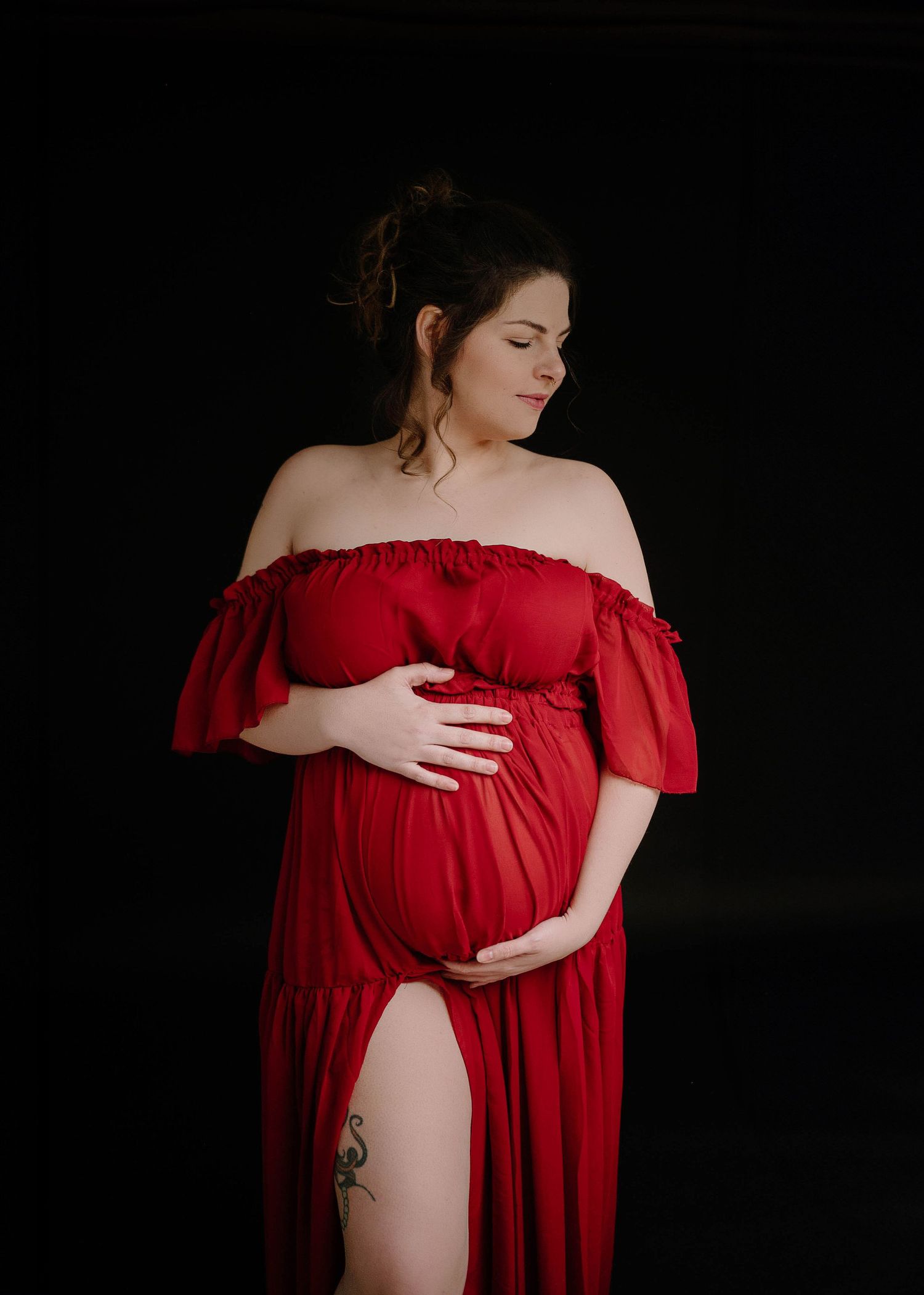 Pregnant woman in red off-shoulder dress holding baby bump against black backdrop.