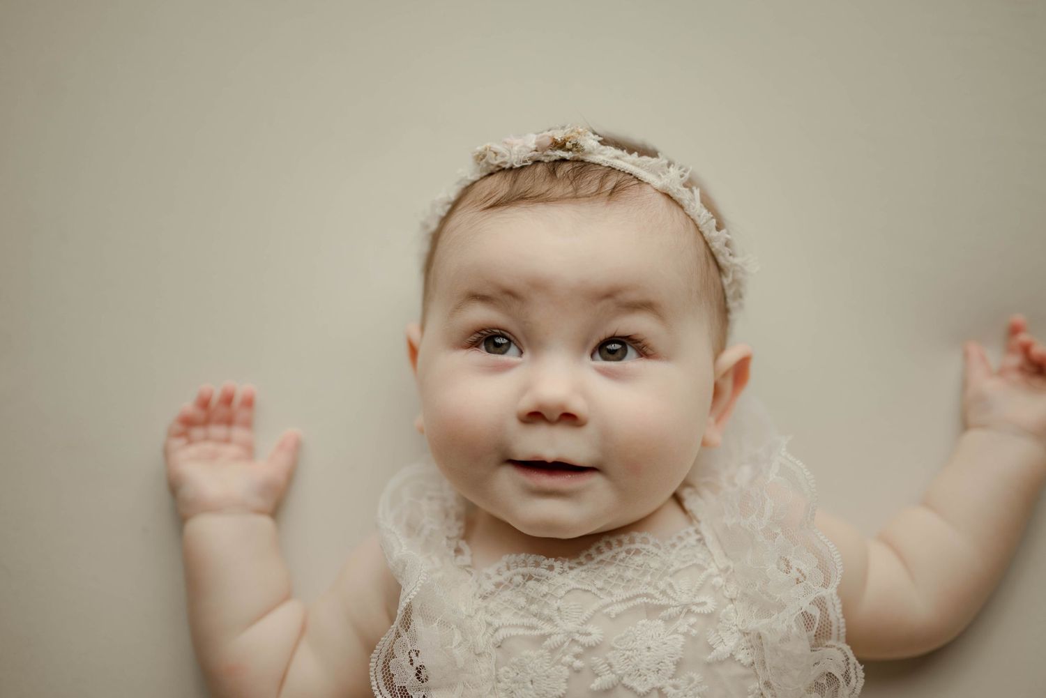 Adorable baby girl in white lace romper and floral headband lying on cream backdrop in studio portrait.