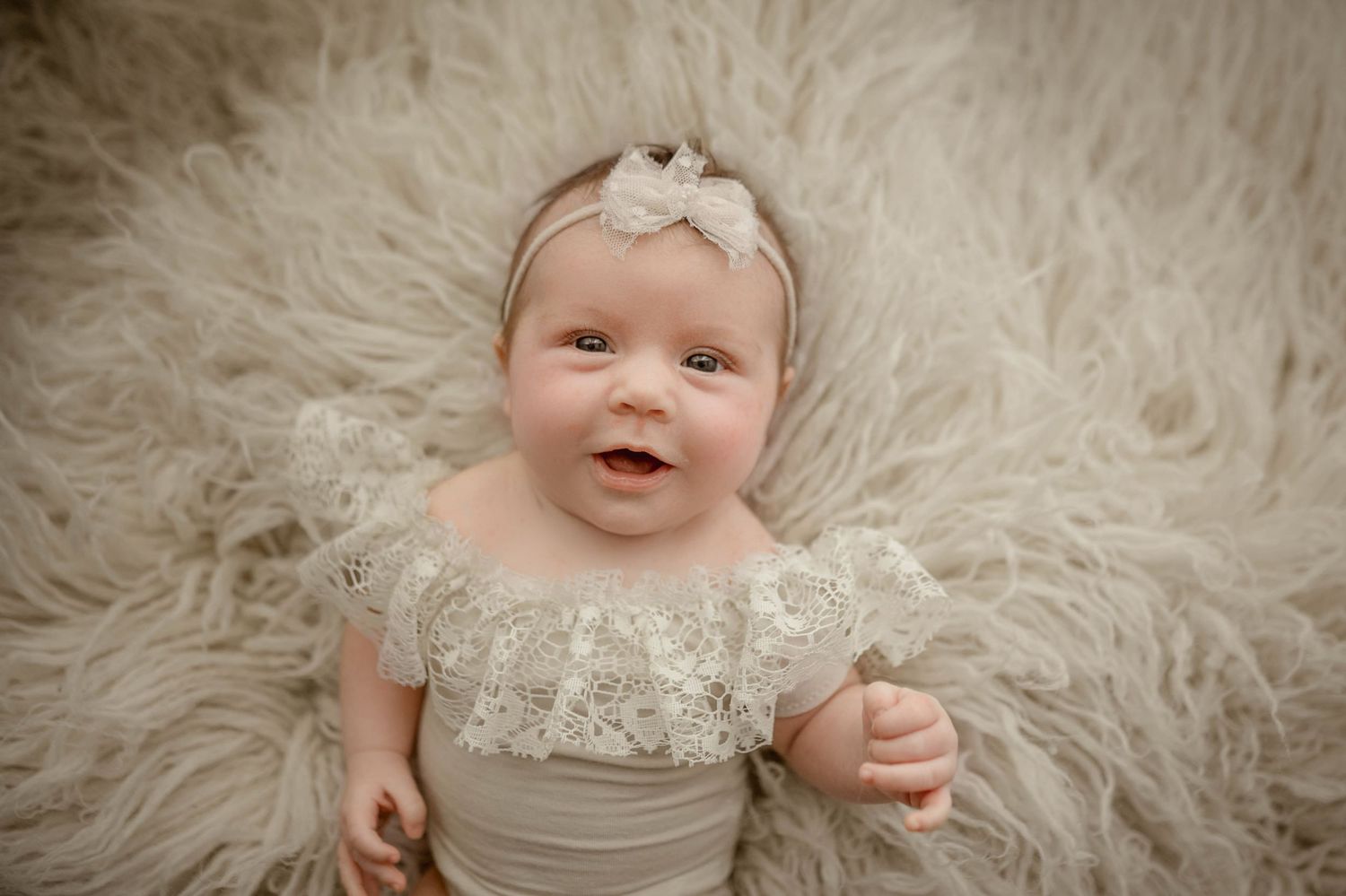 Smiling baby girl in white lace outfit and bow headband lying on fluffy cream fur rug for newborn photo session.