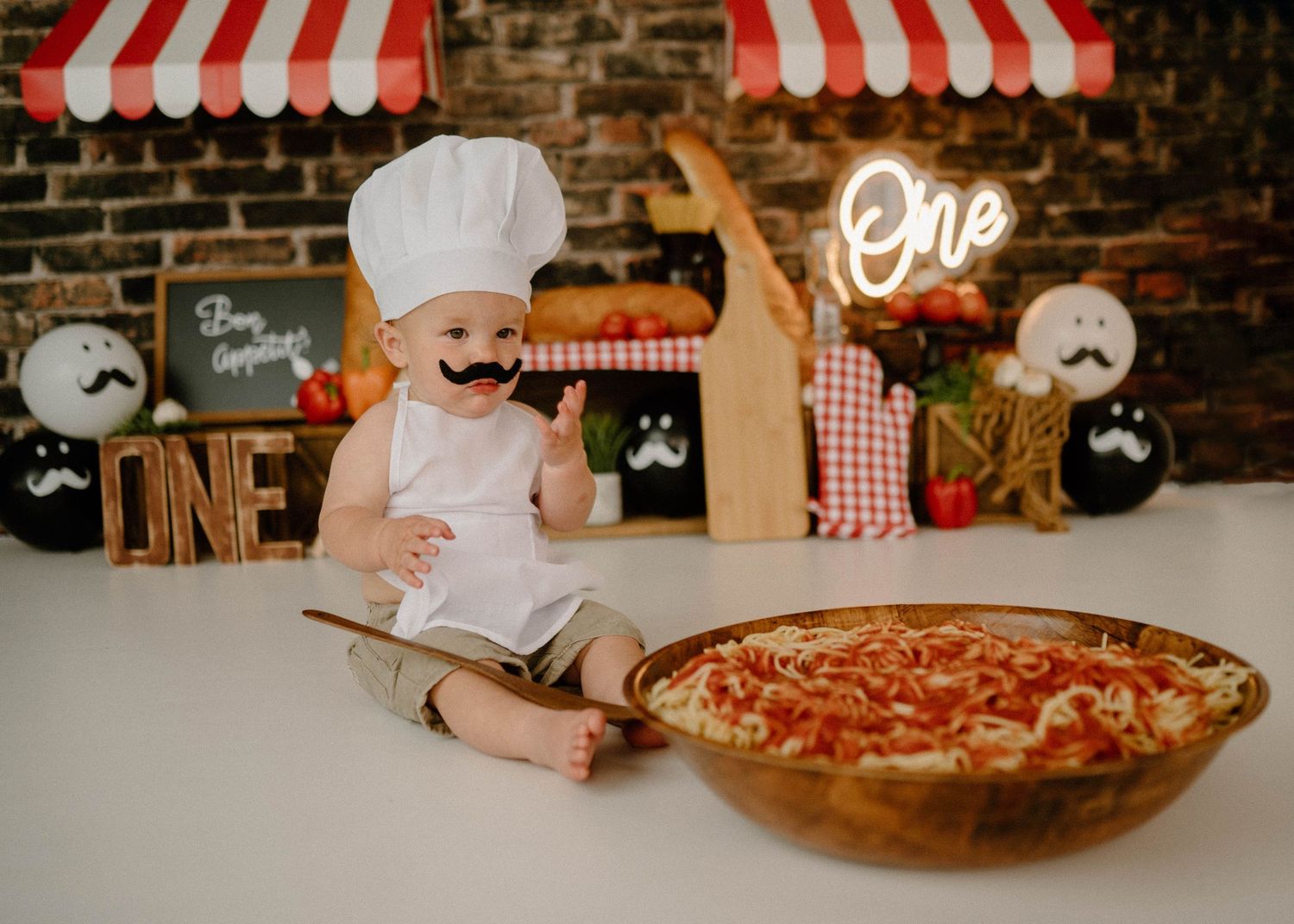 Baby boy in chef hat poses with pizza bowl at Italian-themed first birthday cake smash.