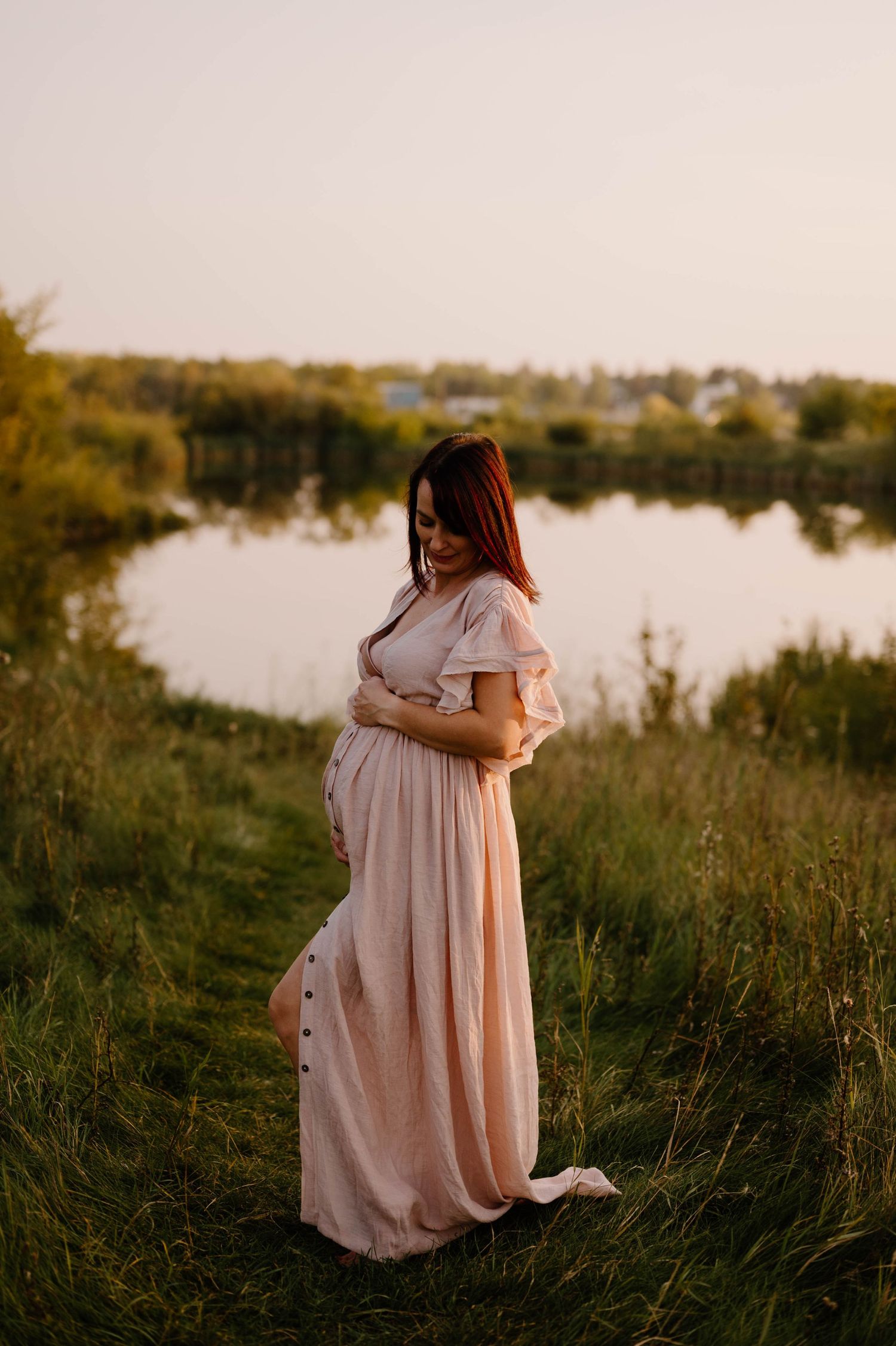 Expectant mother in beige maxi dress cradling baby bump near serene lake Telford.