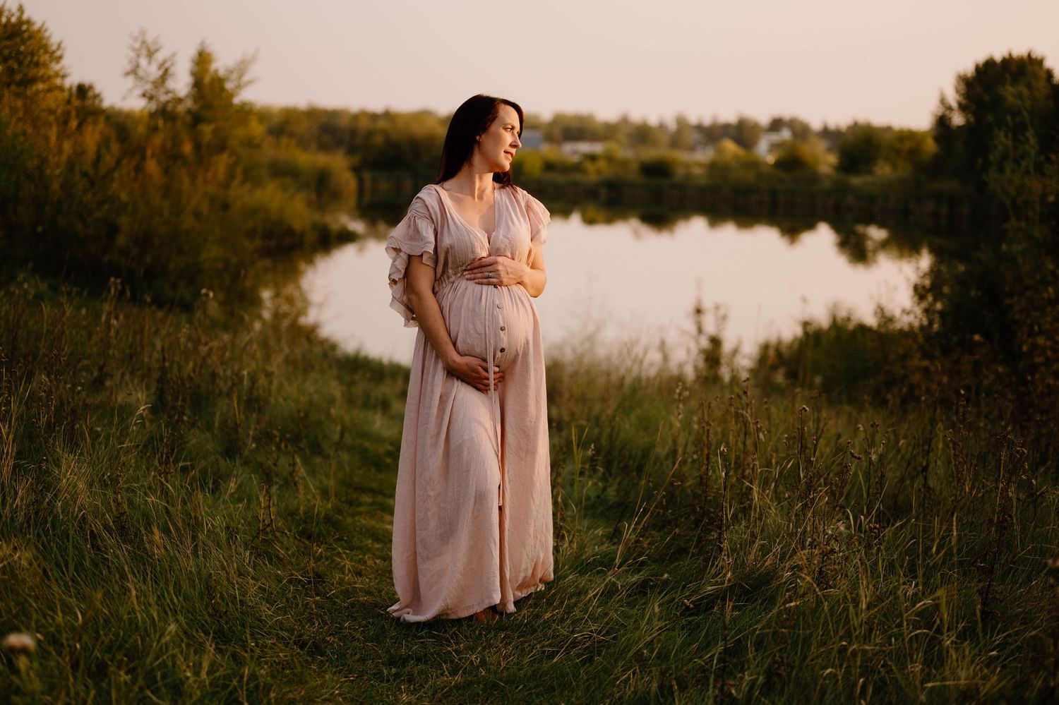 Glowing pregnant woman in pink dress cradles bump while standing in green meadow near Telford Lake at sunset.