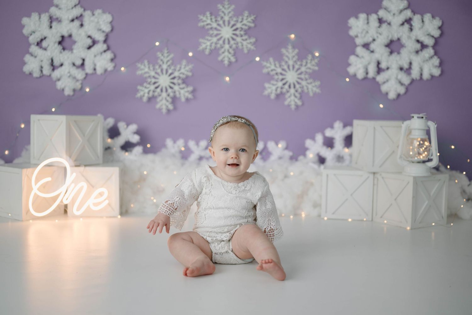 Smiling baby girl in white outfit sitting before a purple snowflake first birthday backdrop.