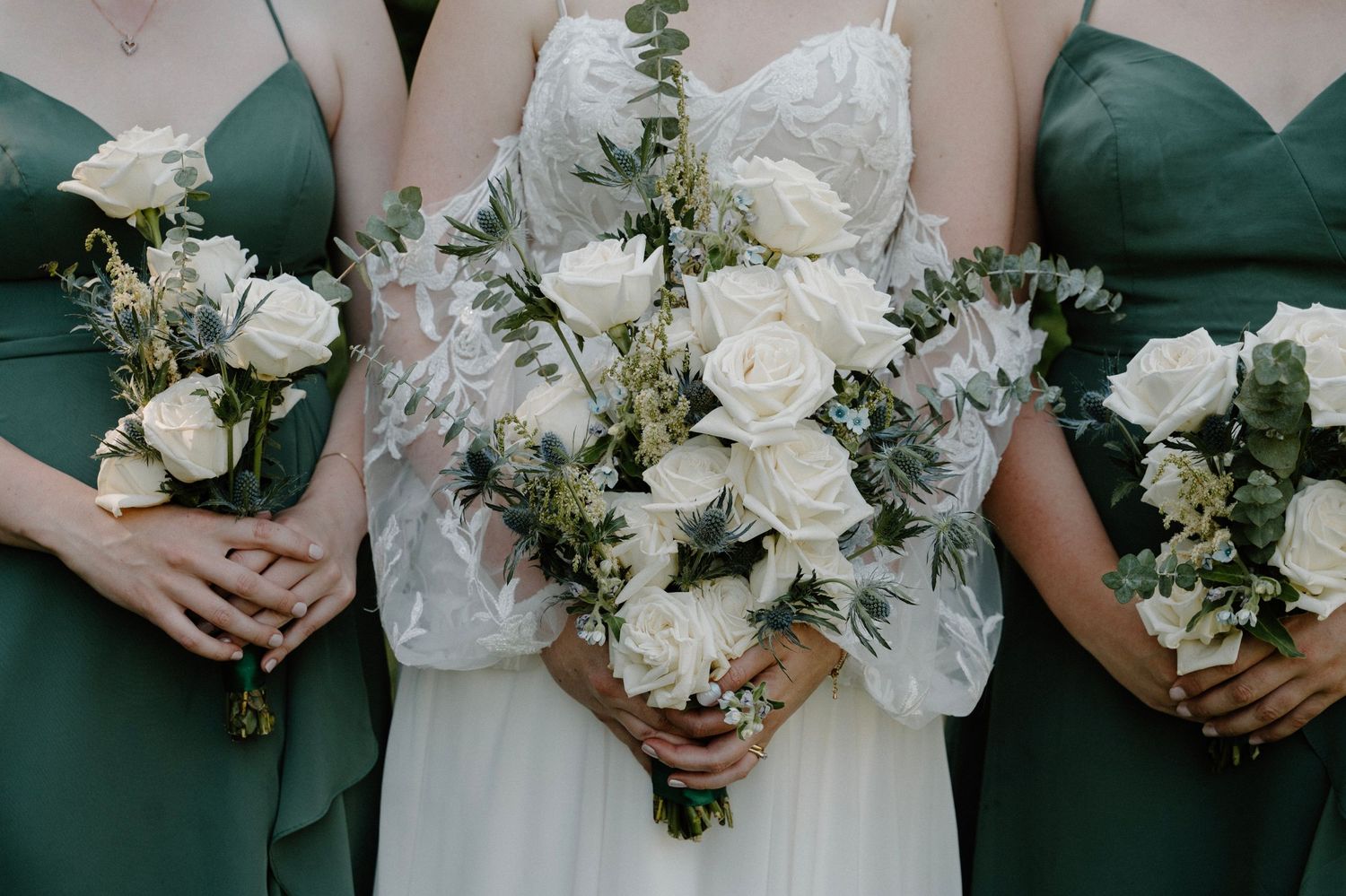 Bride and bridesmaids in sage green dresses hold elegant white rose and eucalyptus bouquets.
