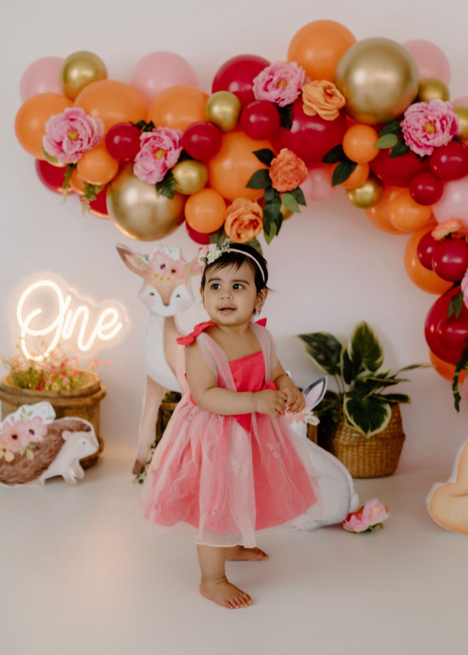 Toddler girl in pink dress standing at first birthday party with balloon arch and woodland decor.