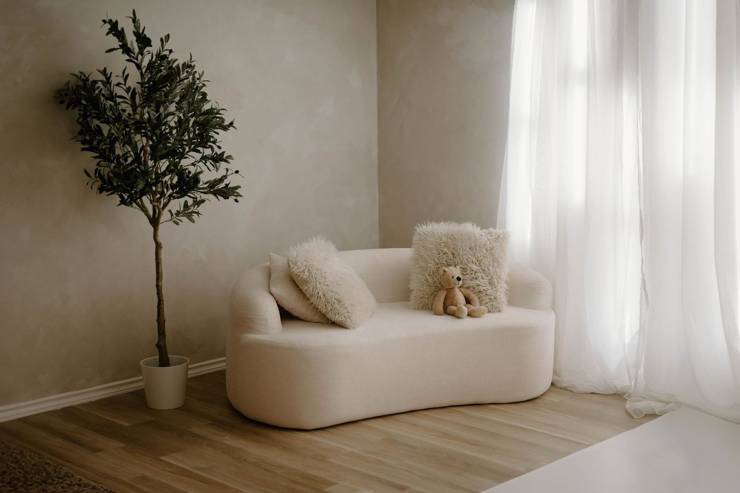 Minimalist living room with a curved white boucle sofa, decorative pillows, potted olive tree, and sheer white curtains.