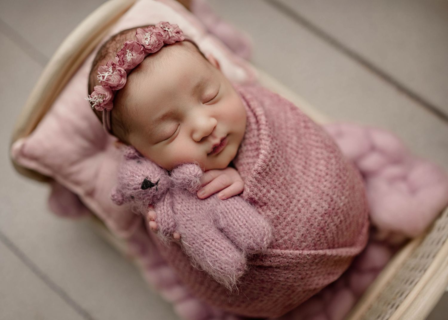 Sleeping newborn baby wrapped in pink knit blanket with floral headband, holding a pink teddy bear in a wooden basket.