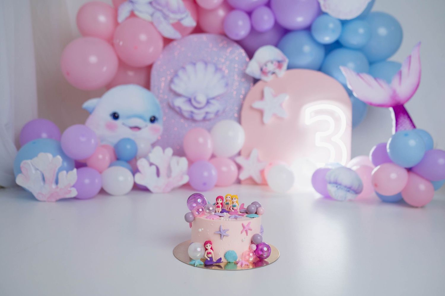 Mermaid themed third birthday cake surrounded by pink purple and blue balloon garland decorations.