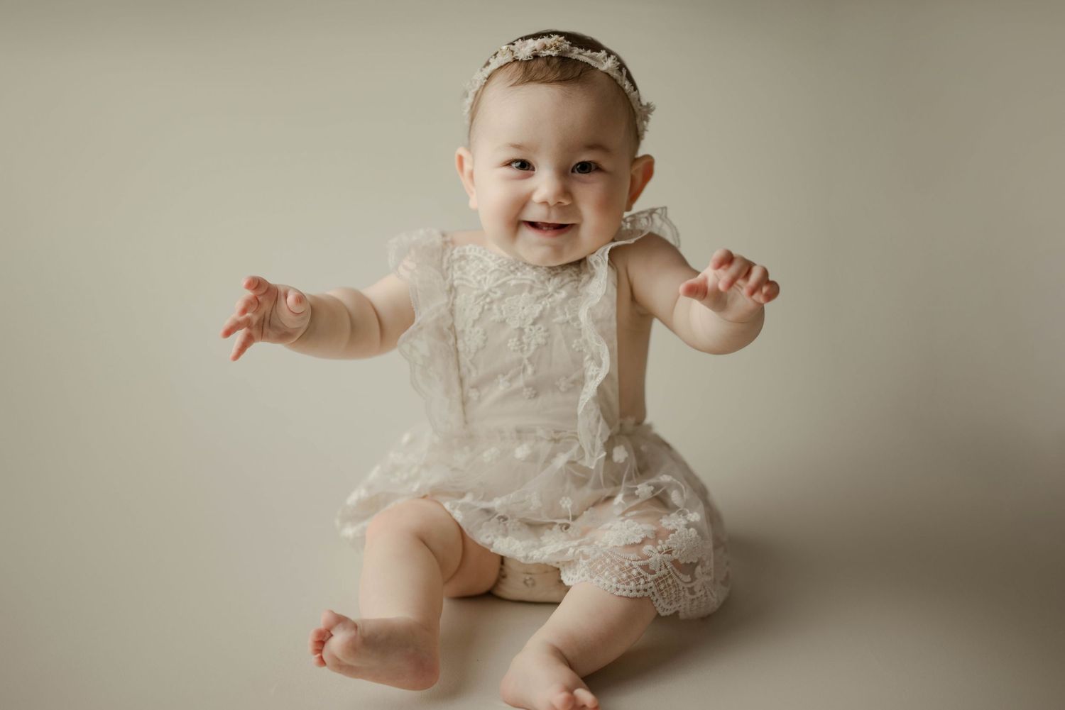 Laughing baby girl in white lace romper and headband sitting up during professional milestone studio portrait.