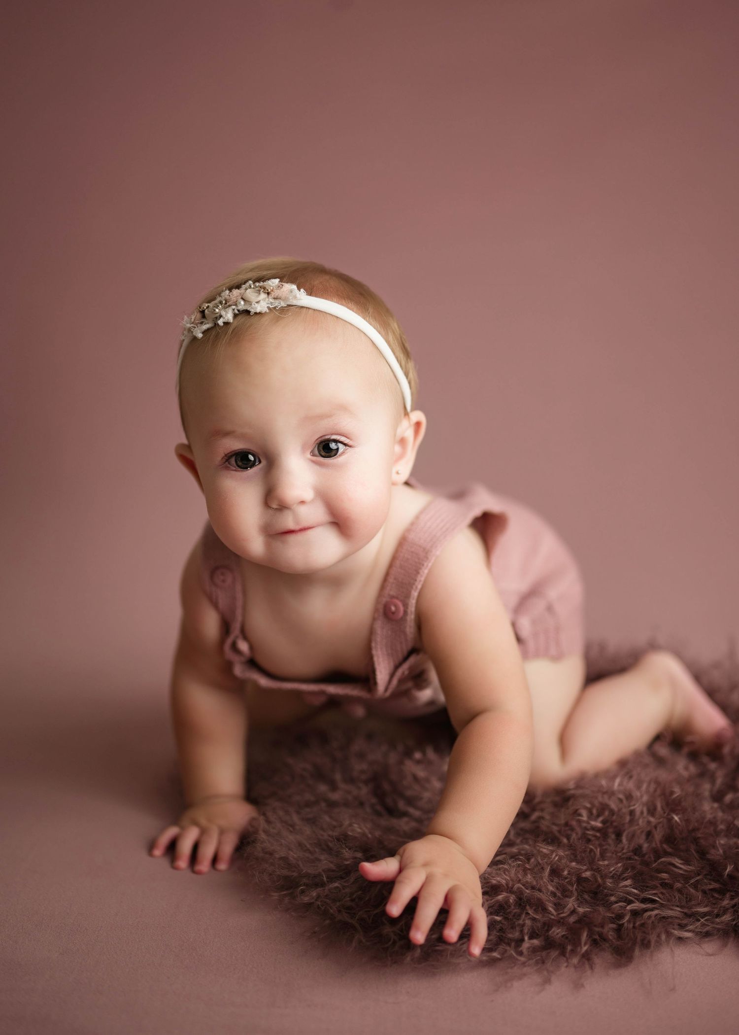 Baby girl in a pink romper and floral headband crawling on a fur rug during a milestone photo session.