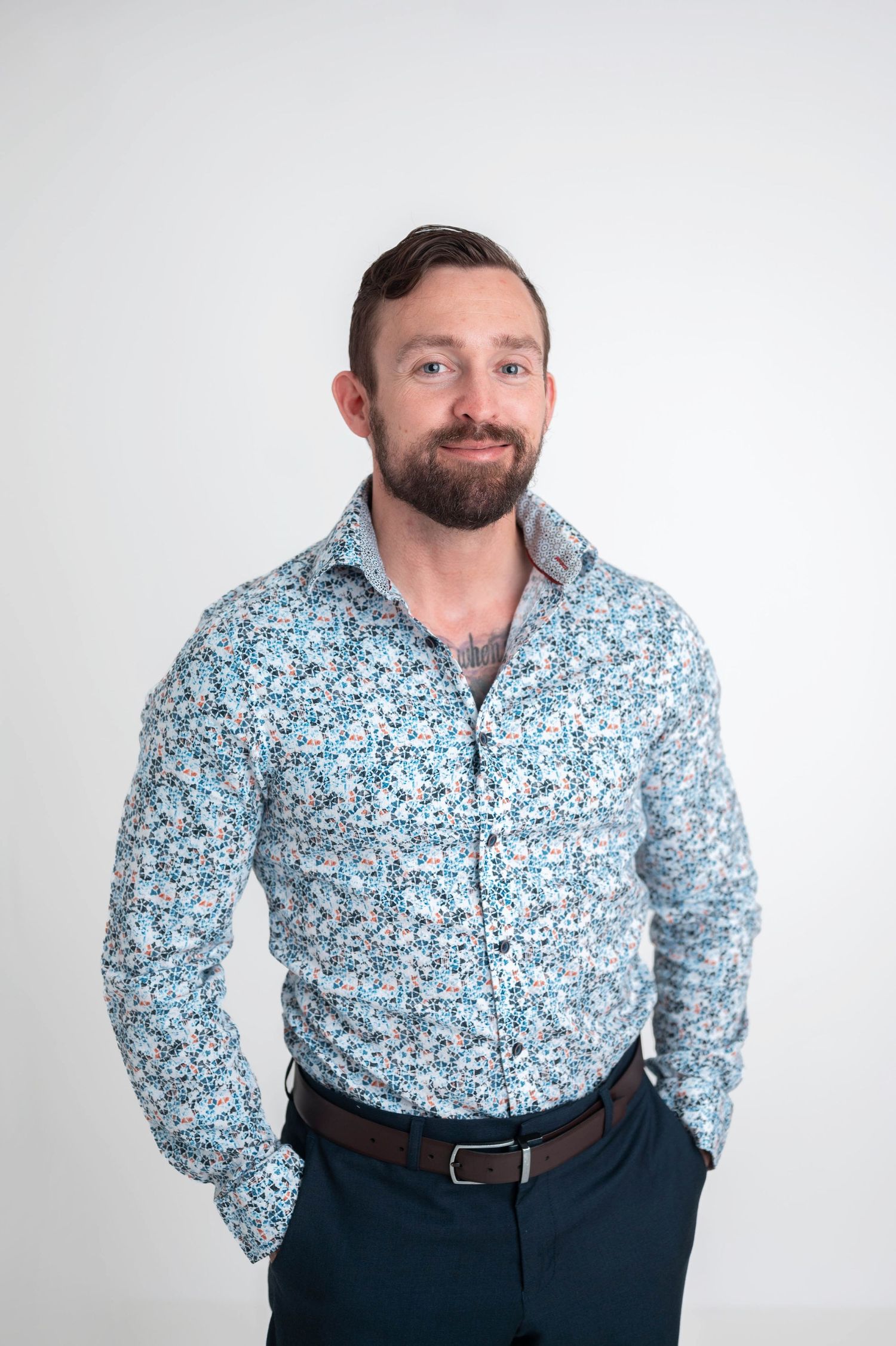 Bearded man smiling in a floral blue button-up shirt and dark pants, posing against a white background.