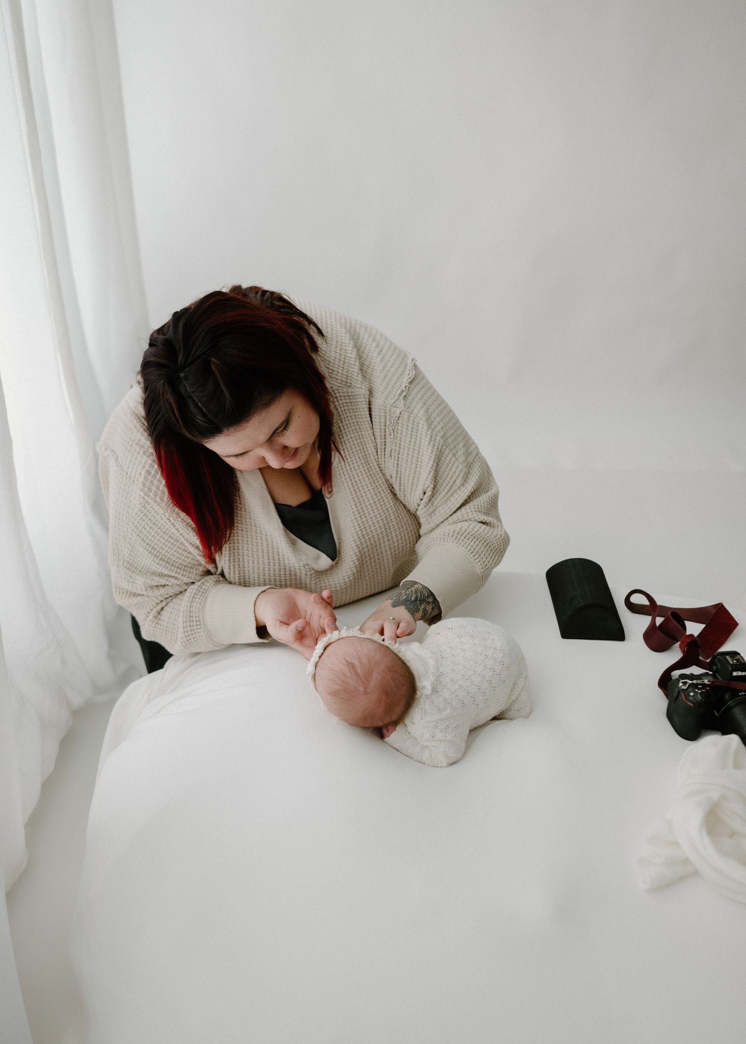 A woman in a cream cardigan gently tends to her sleeping newborn baby wrapped in white on a soft white surface.