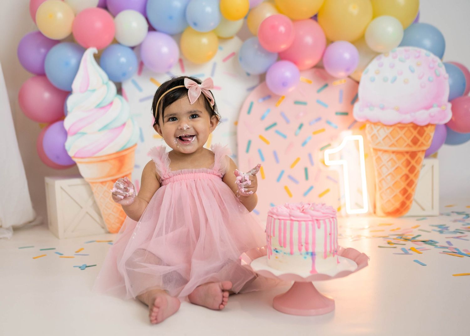 Smiling baby girl in pink dress celebrates first birthday with ice cream themed cake smash backdrop.