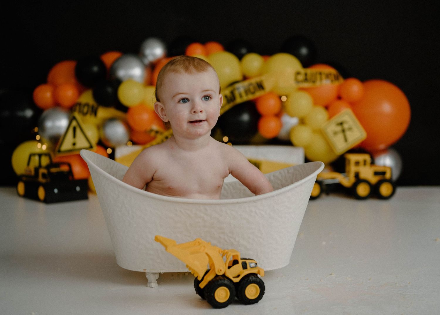 Baby boy sits in white bathtub with yellow toy excavator at construction-themed first birthday setup.