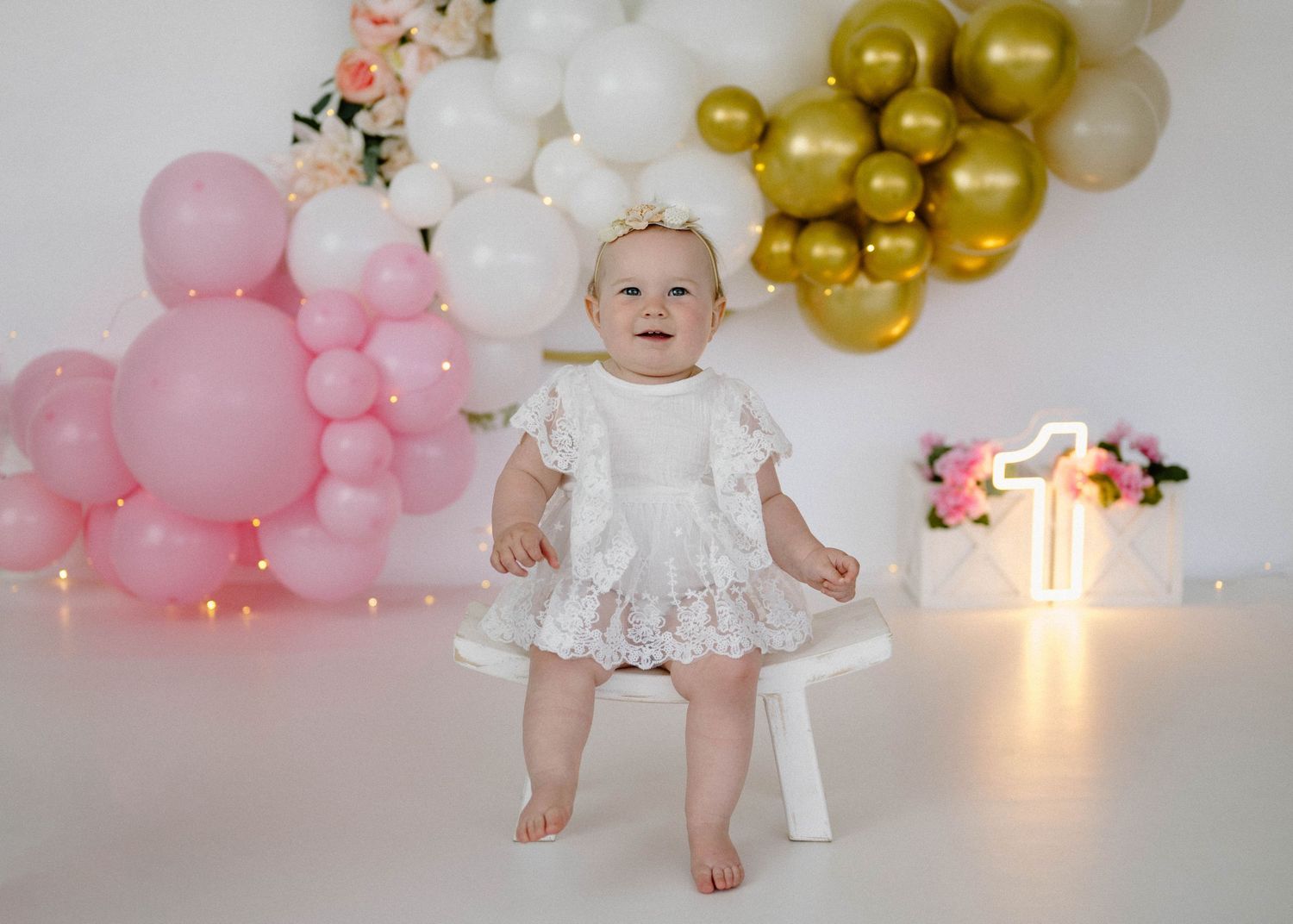 Baby girl in white lace dress stands at bench at first birthday with pink, white, and gold balloon backdrop.
