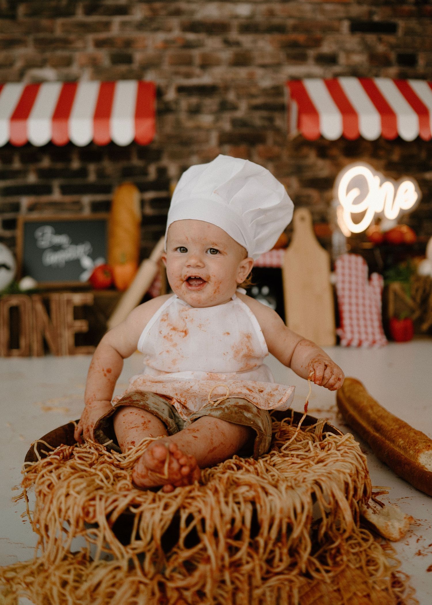 Smiling baby wearing chef hat sits in spaghetti bowl at Italian restaurant themed first birthday shoot.