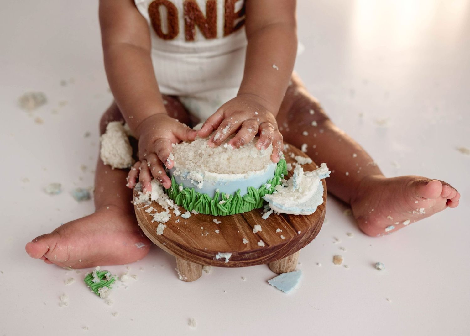 Baby smashing first birthday cake on wooden stand, hands covered in white frosting and green decorations.
