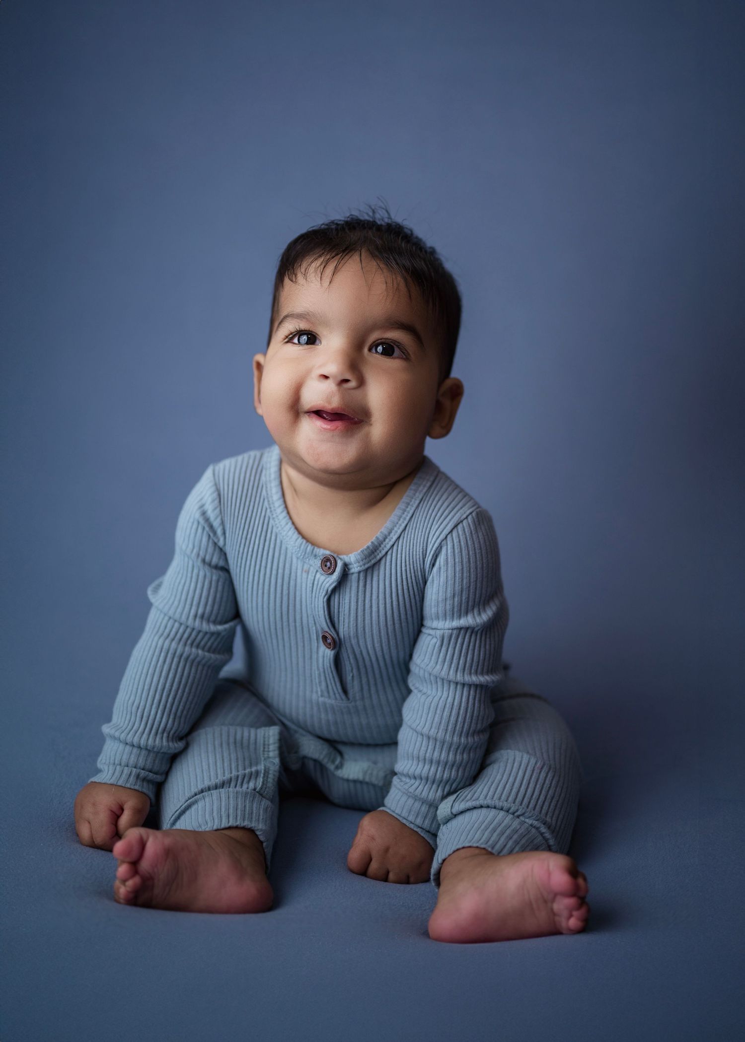 Smiling baby in blue ribbed romper sitting on blue background.