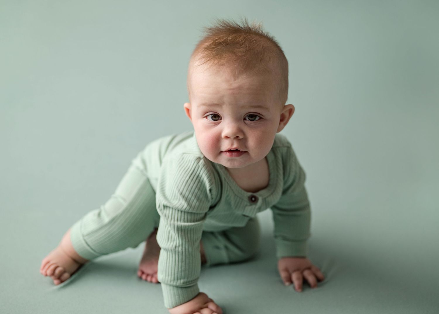 Red-haired baby in mint green ribbed romper crawling on teal floor during professional studio photo session.