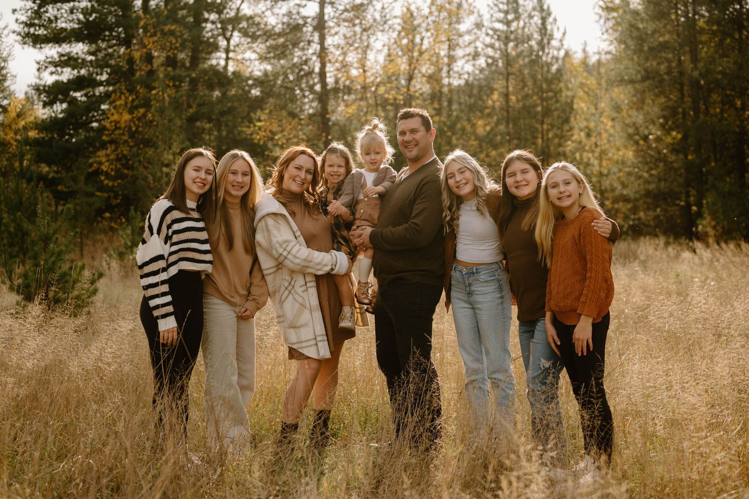 Large family of nine smiling together in a sunlit autumn forest clearing.