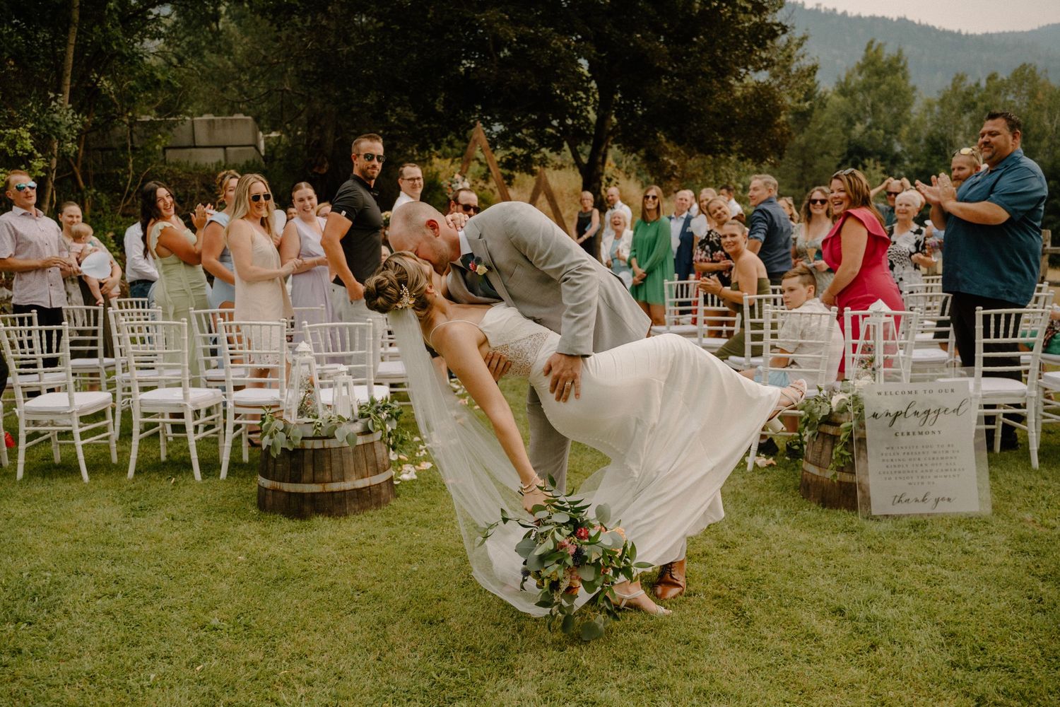 Groom dipping bride for a romantic kiss on the lawn surrounded by cheering wedding guests.