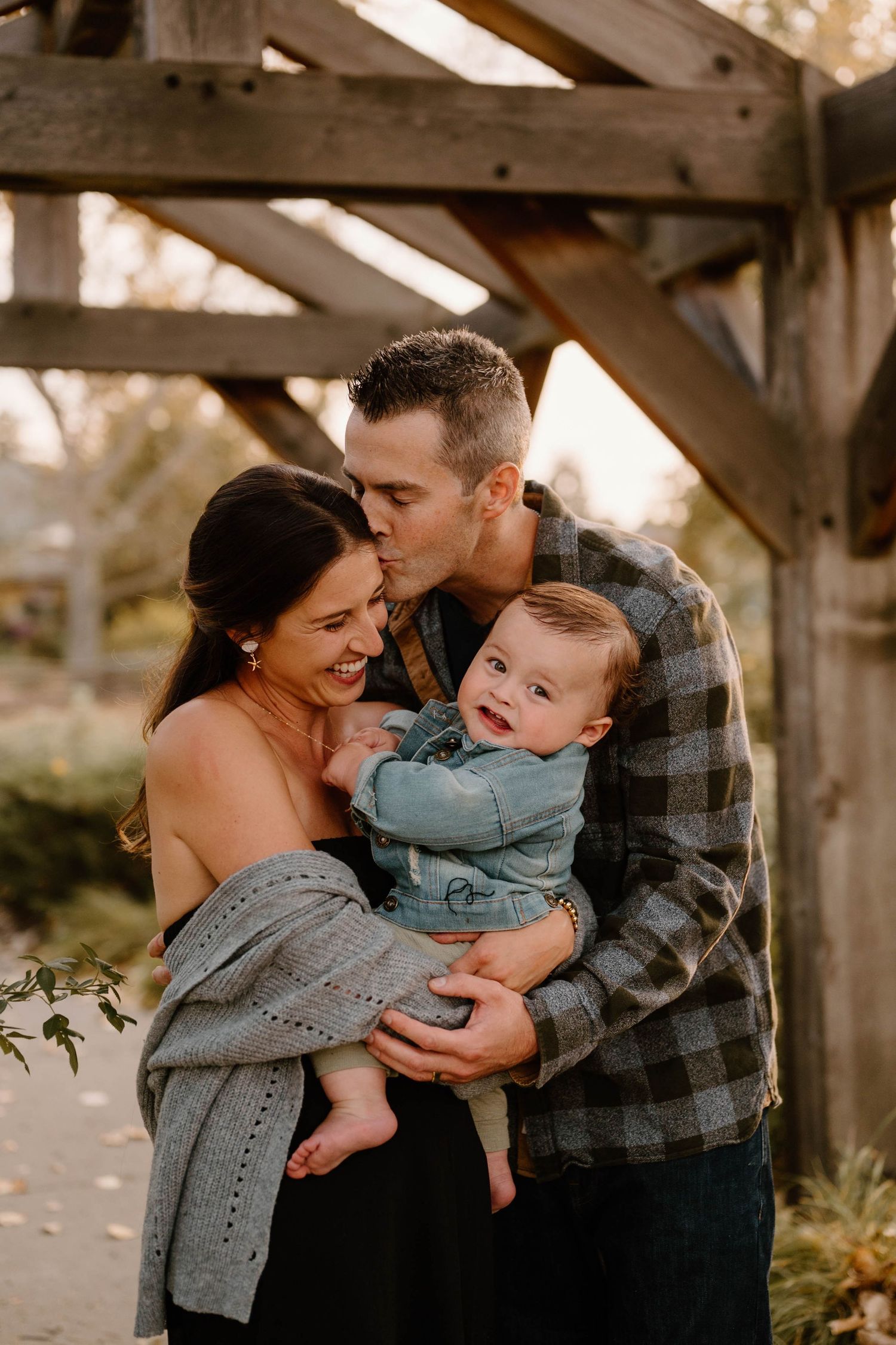 Loving father kisses mother's head as couple shares joyful moment holding laughing baby outdoors.
