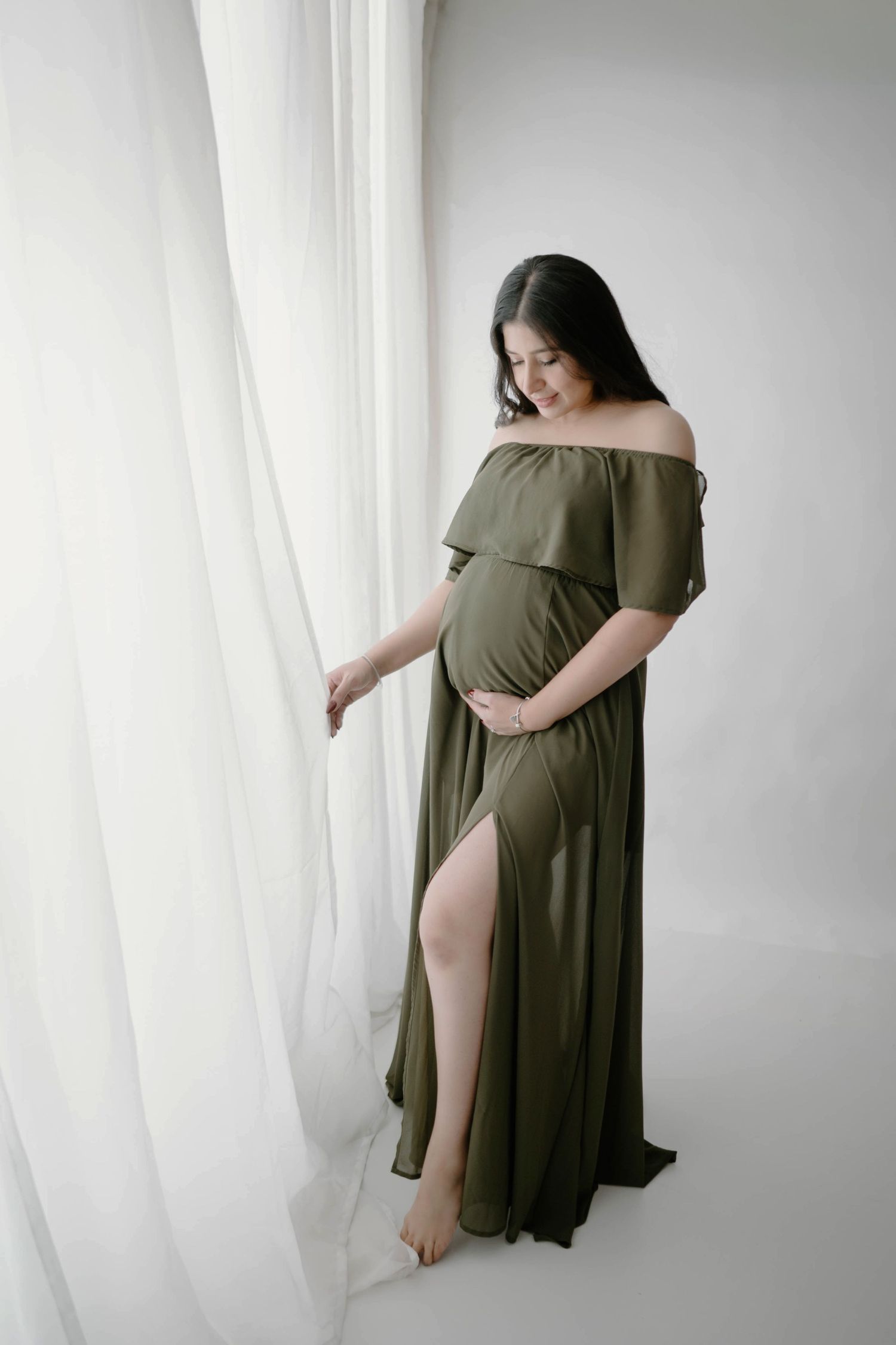 Pregnant woman in olive off-shoulder gown standing by sheer white curtains in bright Leduc studio.