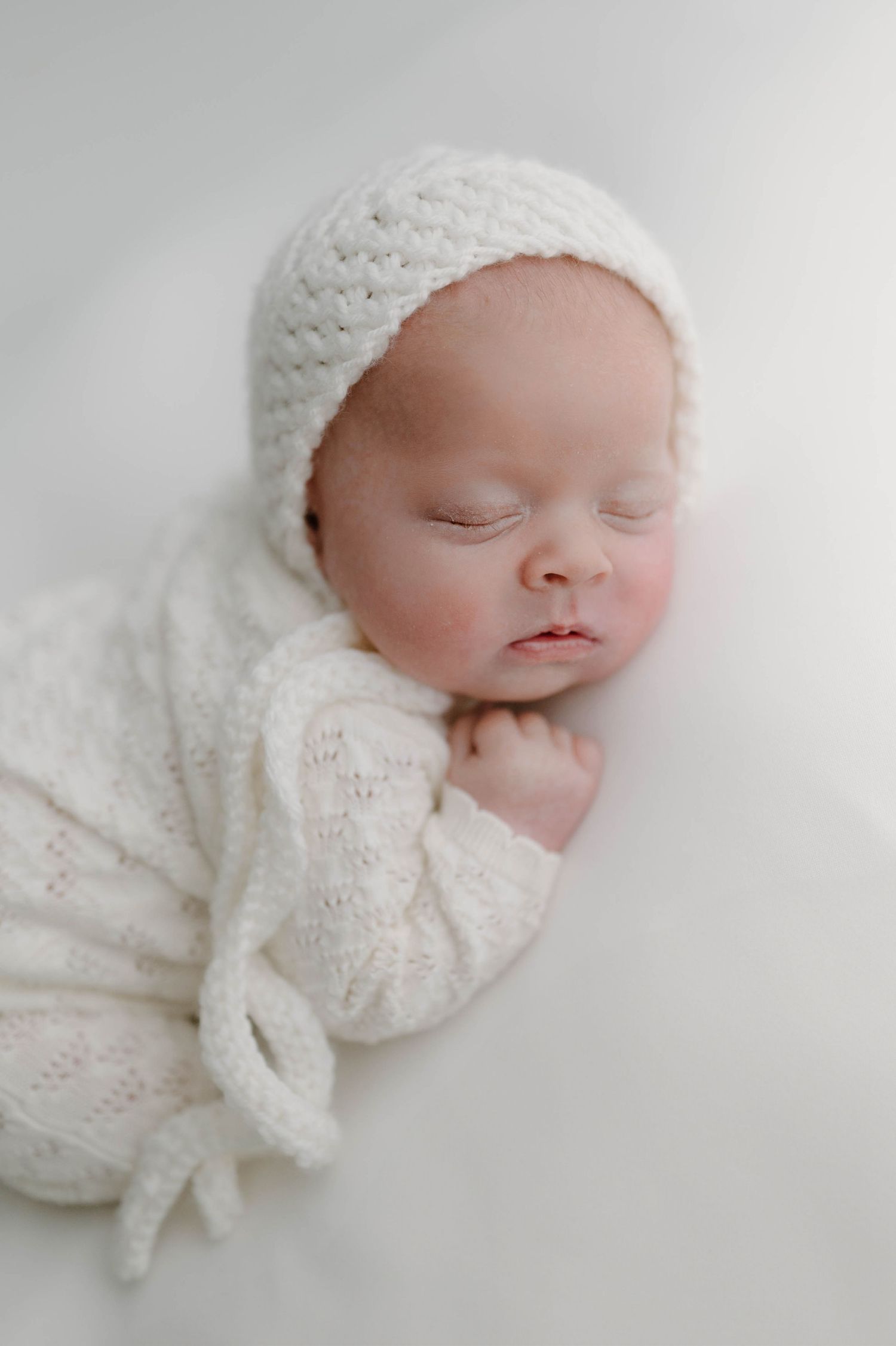 A peaceful newborn baby sleeps on a white blanket with a matching bonnet.