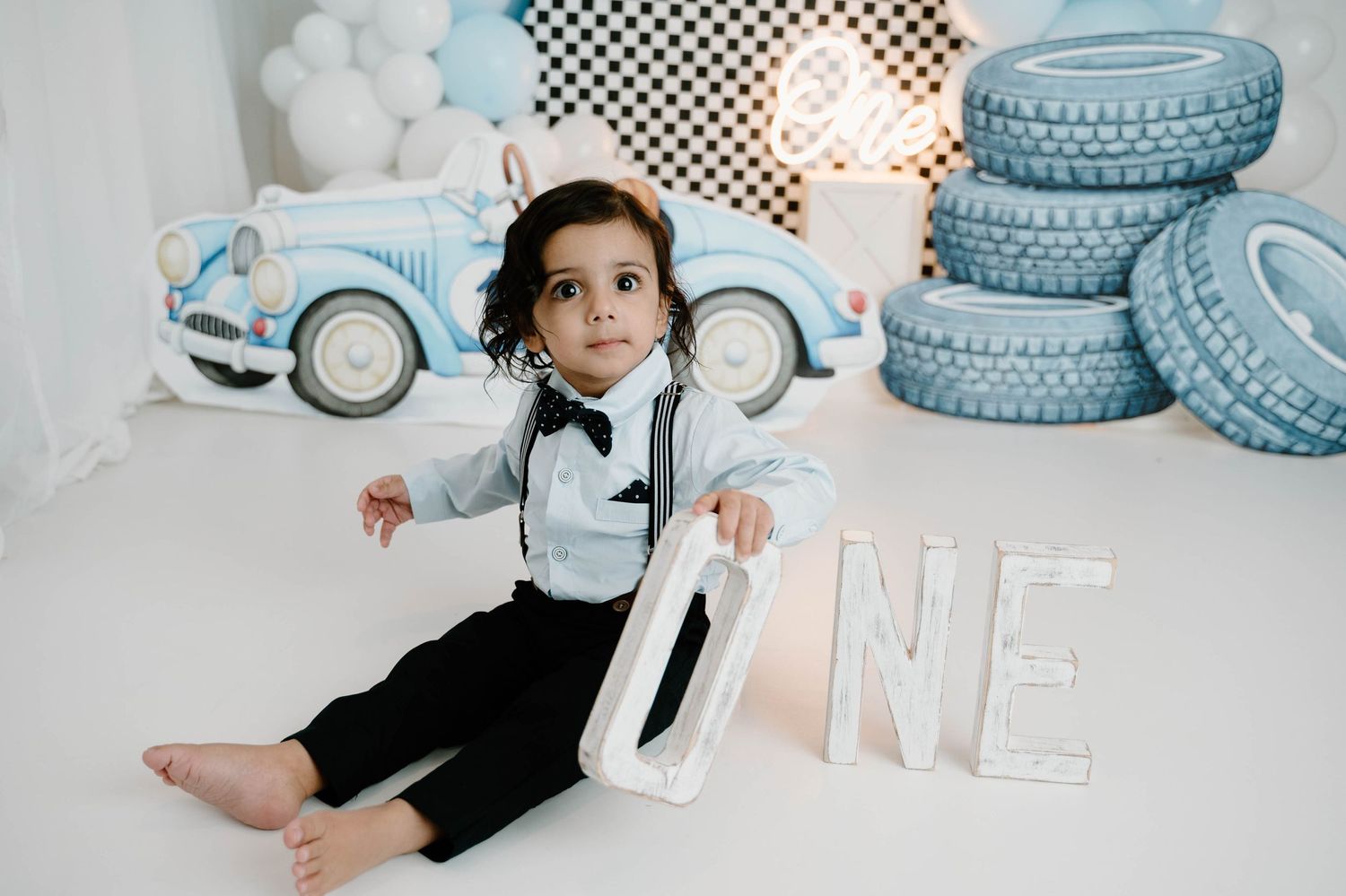 A toddler boy in suspenders and bow tie sits beside large ONE letters at a vintage blue race car first birthday photo session.
