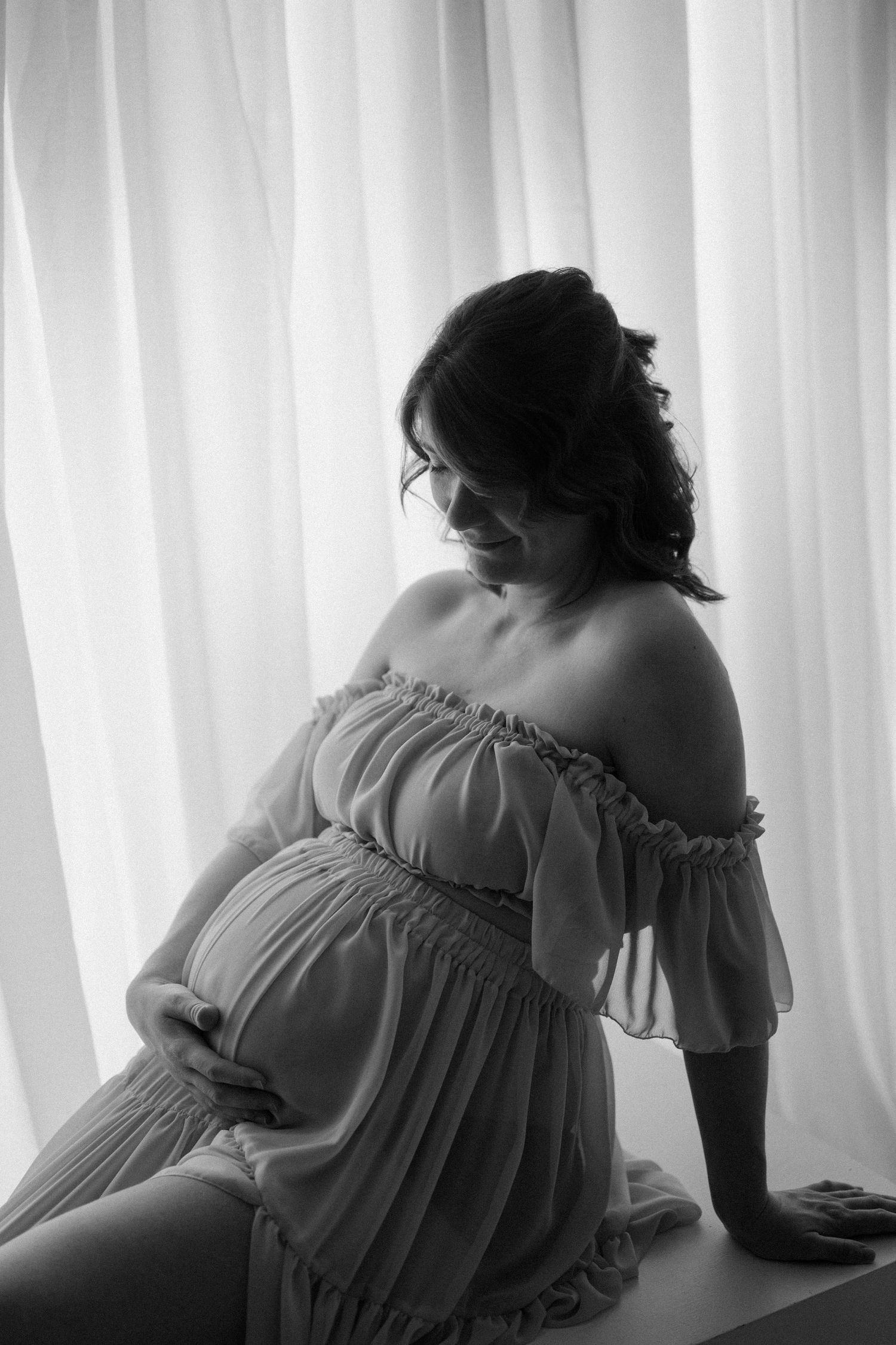 Black and white maternity photo of pregnant woman in off-shoulder dress cradling baby bump by sheer curtains.
