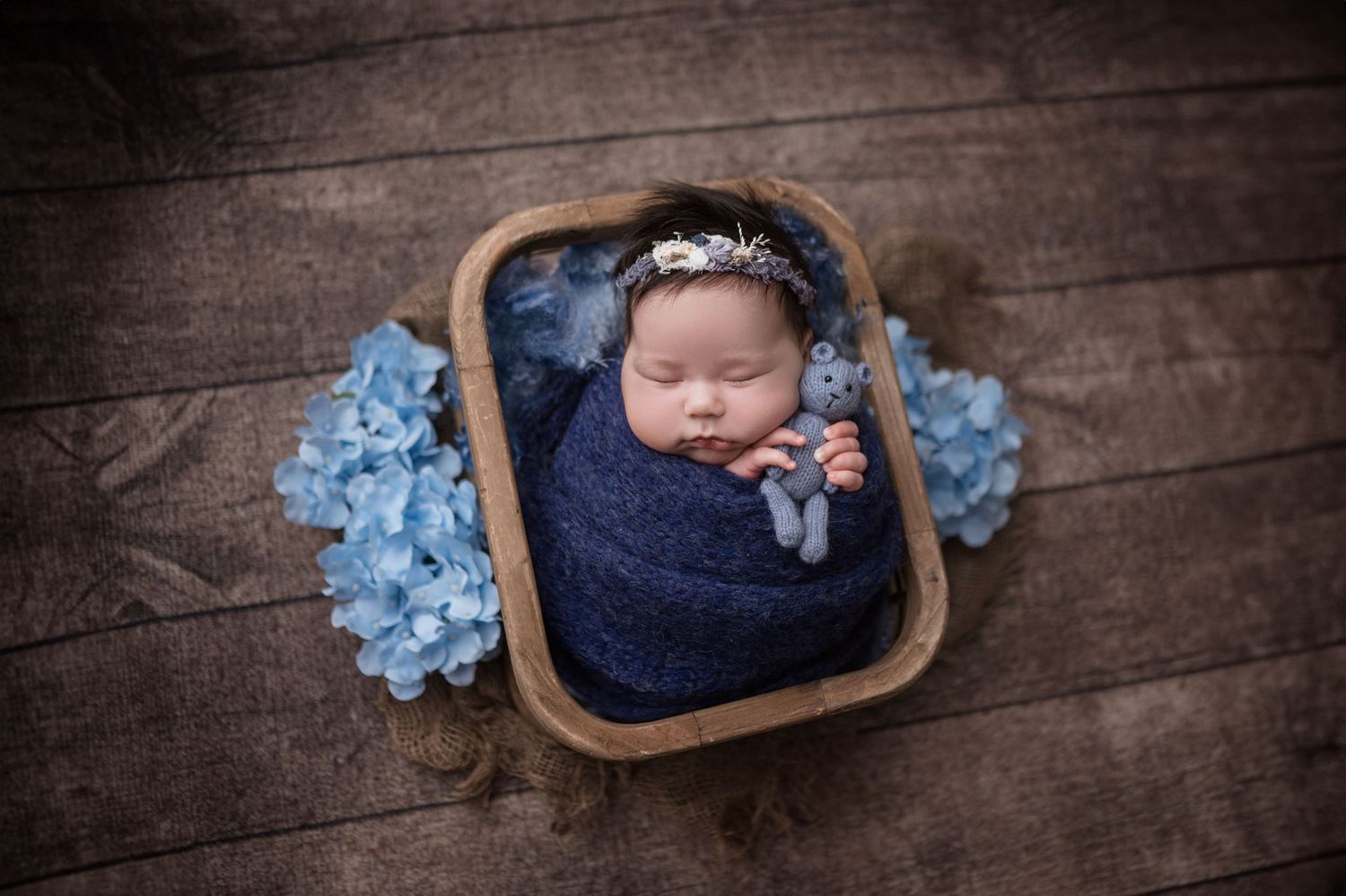Newborn baby swaddled in navy blue wrap, sleeping in wooden bowl with blue hydrangeas and floral headband.